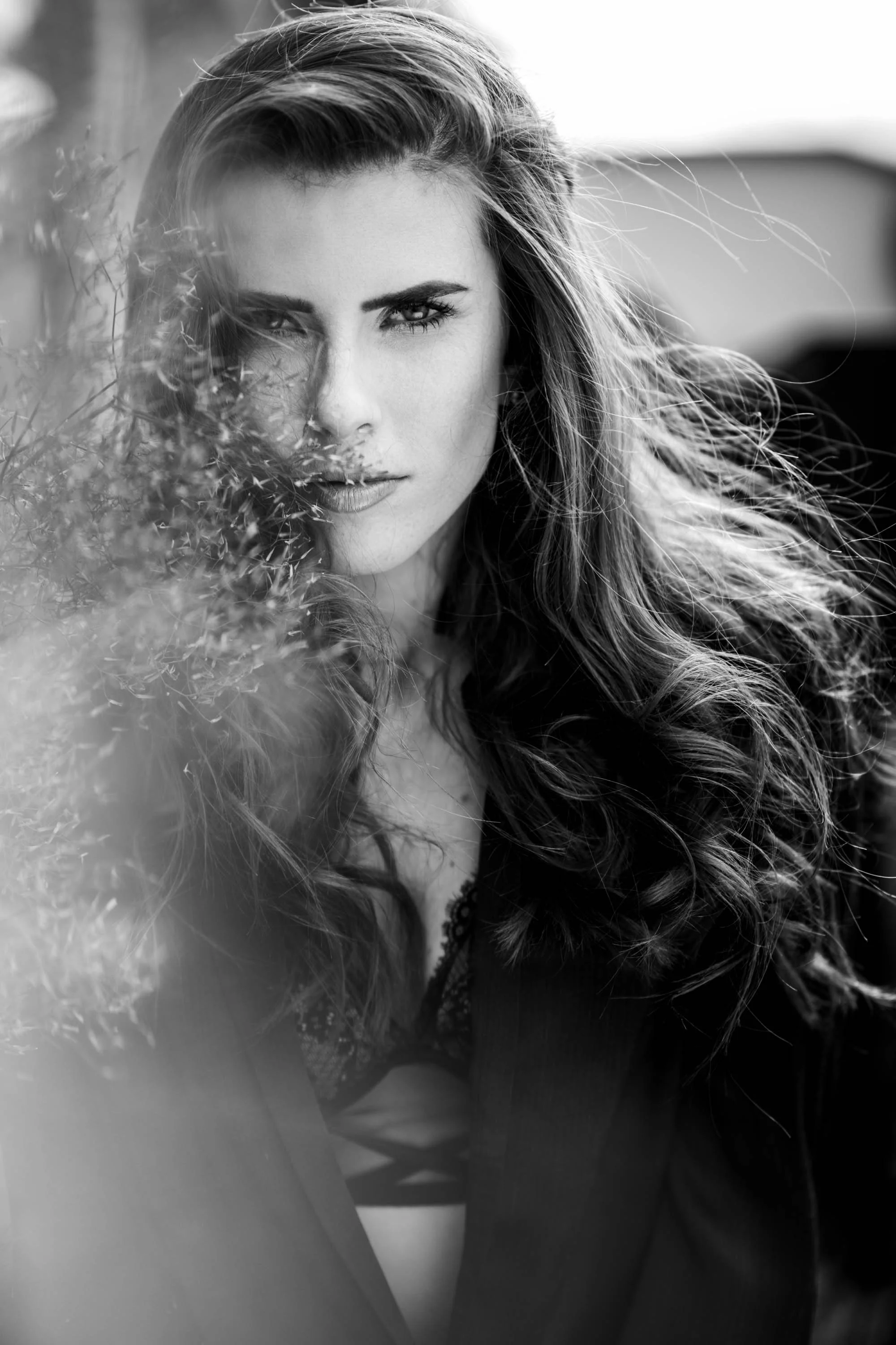 Schwarz-weiß-Fotografie einer Frau mit langen, lockigen Haaren, die direkt in die Kamera schaut, teilweise durch Blätter verdeckt.