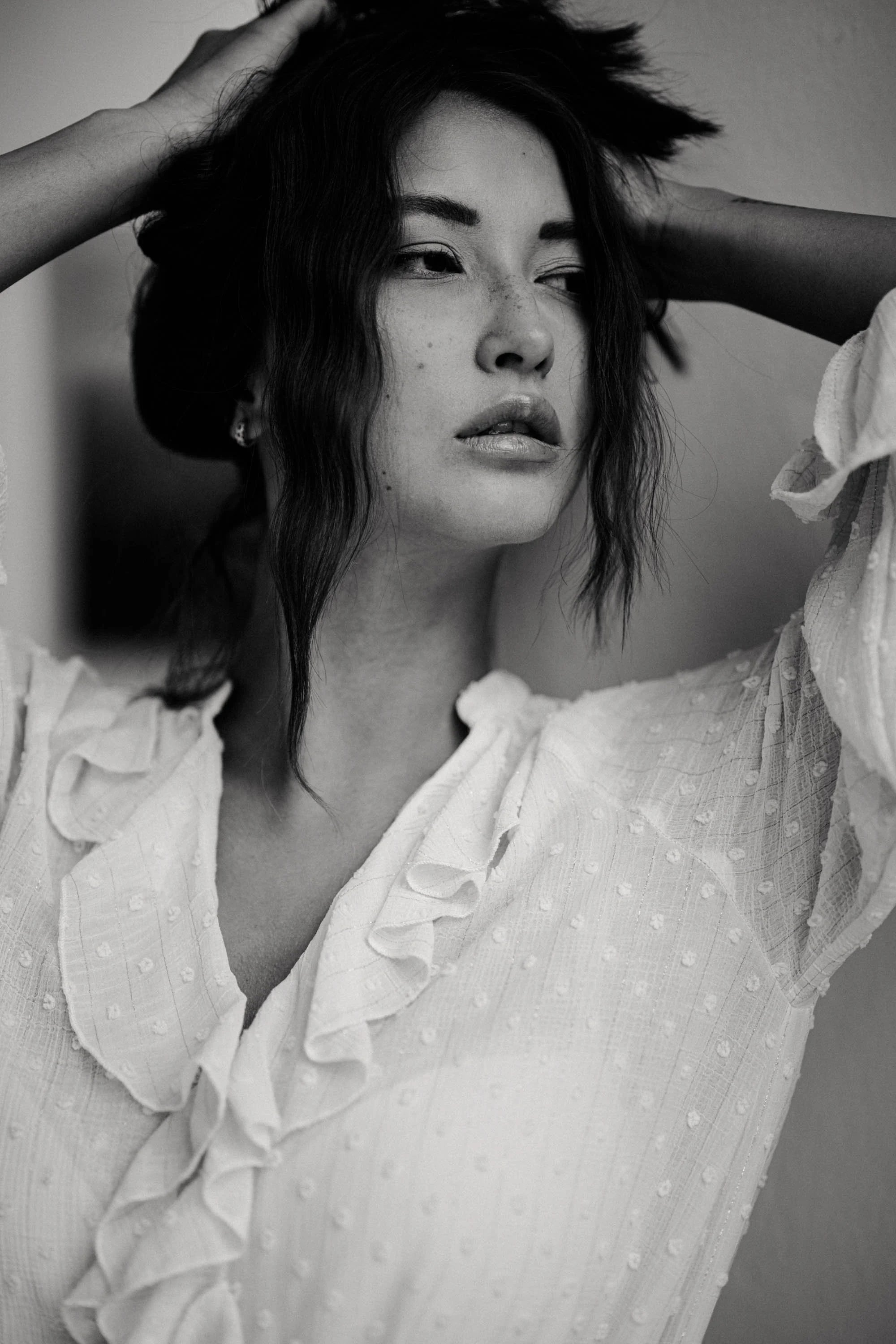 Schwarz-weiß Porträt einer jungen Frau mit dunklen, gewellten Haaren, die eine weiße Bluse trägt, die über die Schultern fällt, während sie den Kopf leicht zur Seite neigt und die Augen halb schließt