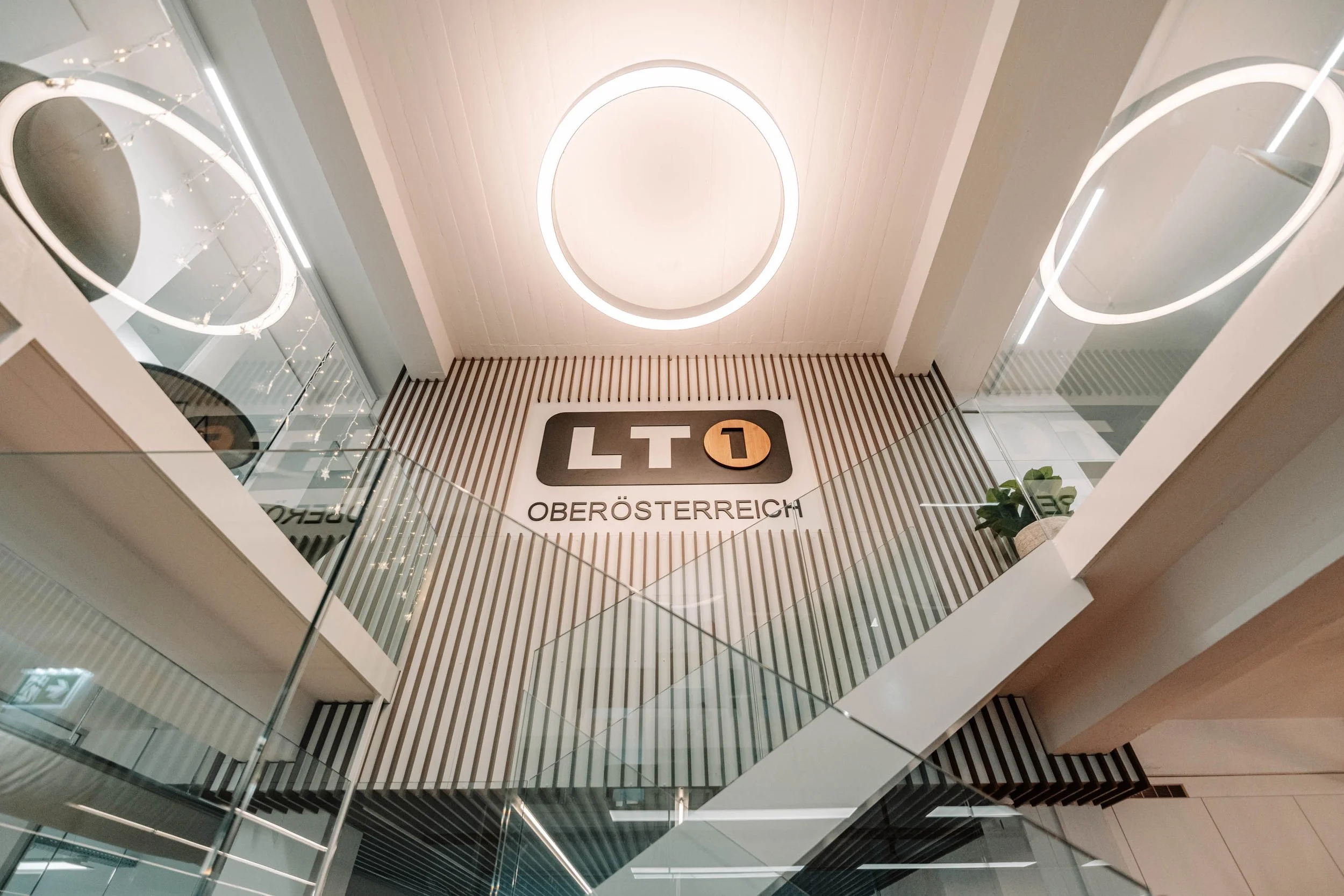 Innenaufnahme eines modernen Gebäudes mit mehreren Stockwerken, Blick nach oben auf eine weiße Decke mit runden Lichtinstallationen, die Wand hat vertikale Holzleisten, und es gibt ein großes Schild mit dem Logo 'LT1' und der Aufschrift 'OBERÖSTERREI