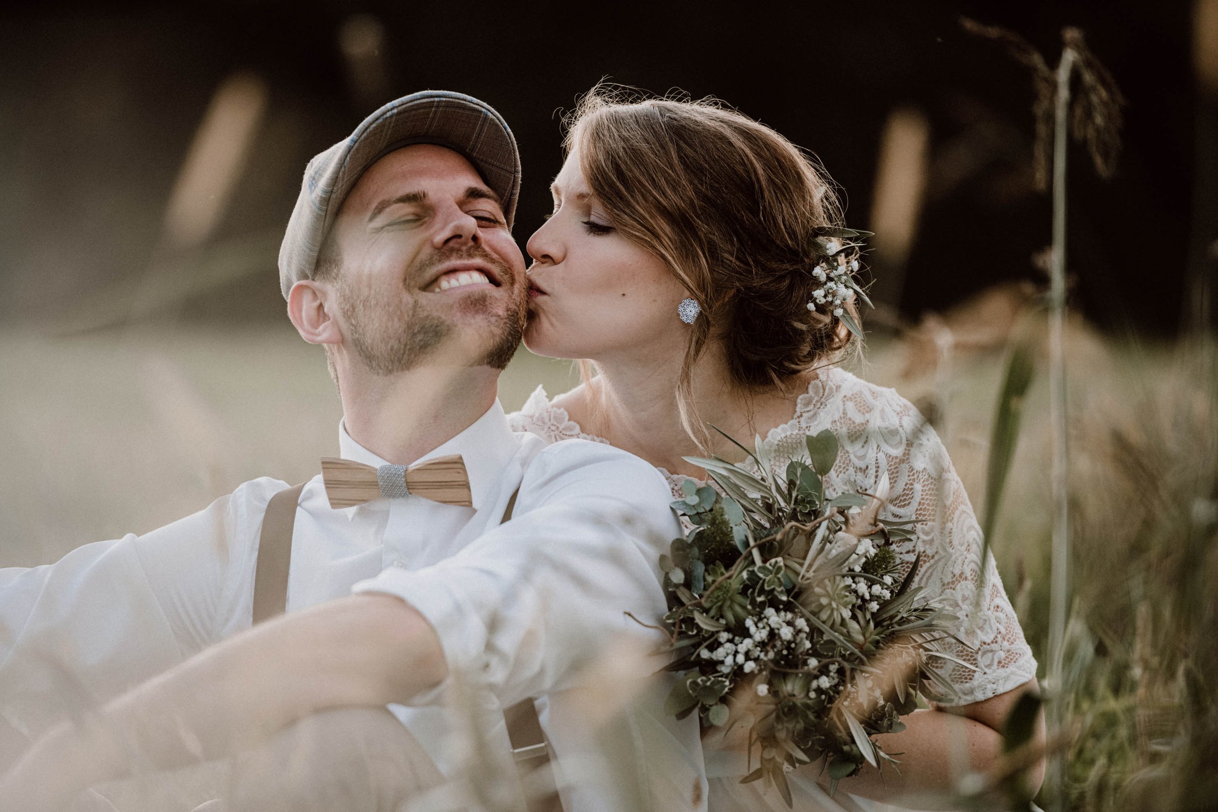 Ein glückliches Paar an ihrer Hochzeit, die Frau küsst den Mann auf die Wange, während er lächelnd im Freien sitzt. Sie trägt ein weißes Spitzenkleid und der Mann trägt Hemd mit Fliege.