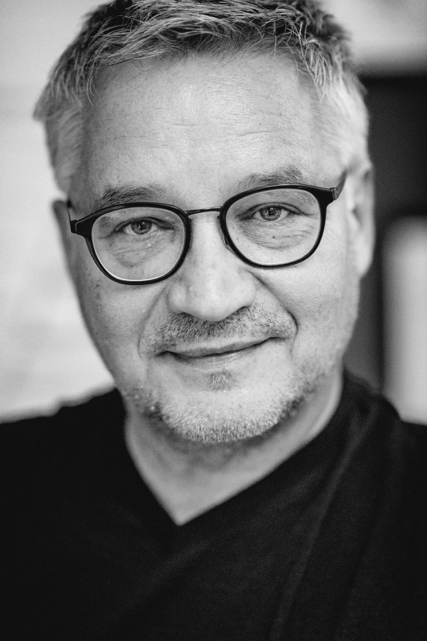 Schwarz-weiß Portrait eines Mannes mit Brille, kurze Haare und leichter Bart, lächelnd mit einem freundlichen Ausdruck.