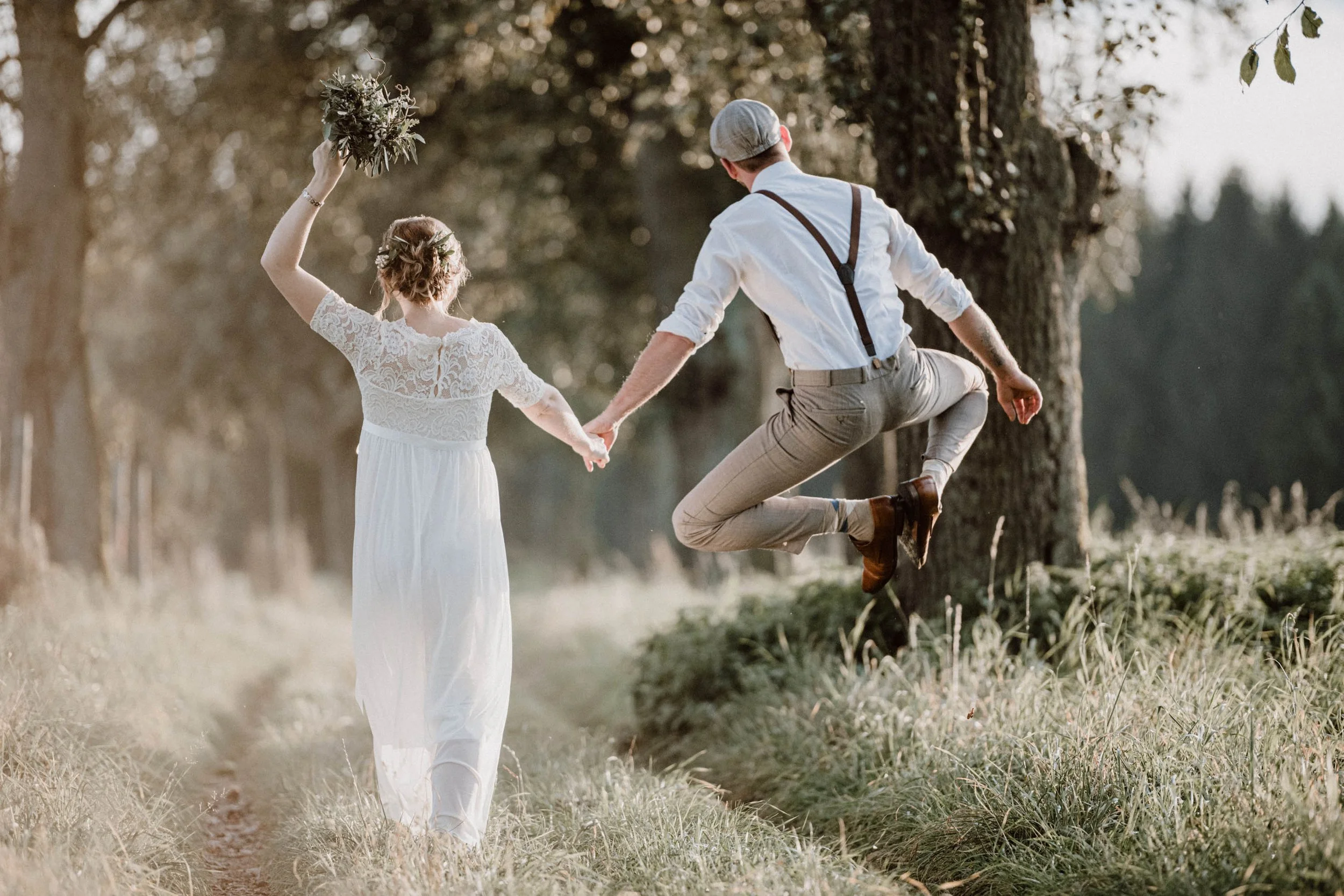 Ein heiratendes Paar läuft Hand in Hand durch ein Feld, das Mann springt in die Luft, die Frau trägt ein langes weißes Kleid und hält einen Blumenstrauß. Es ist ein sonniger Tag, umgeben von Bäumen.