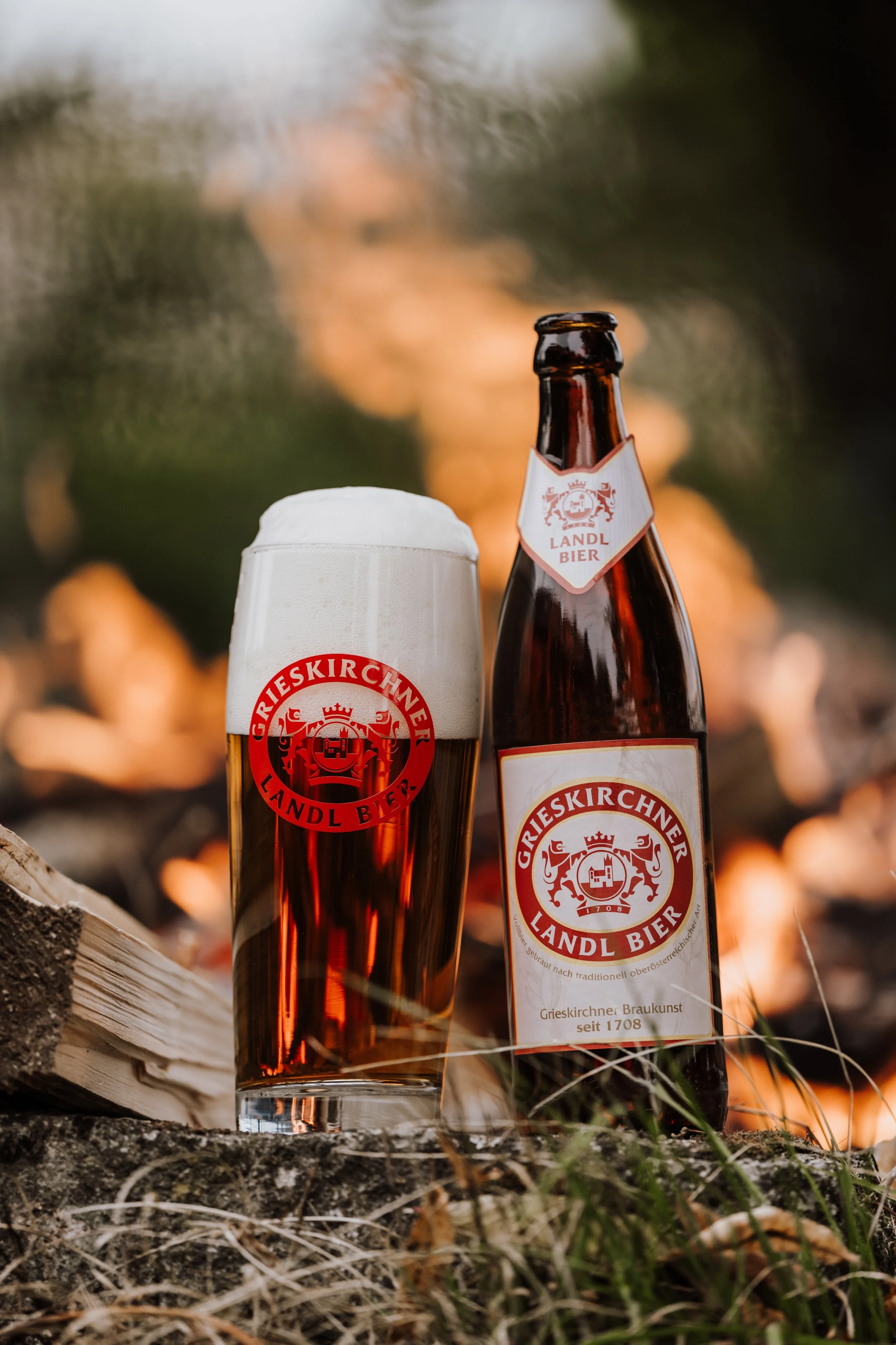 Ein Glas und eine Flasche Bier auf einem Holzstamm im Freien, mit einem Lagerfeuer im Hintergrund.