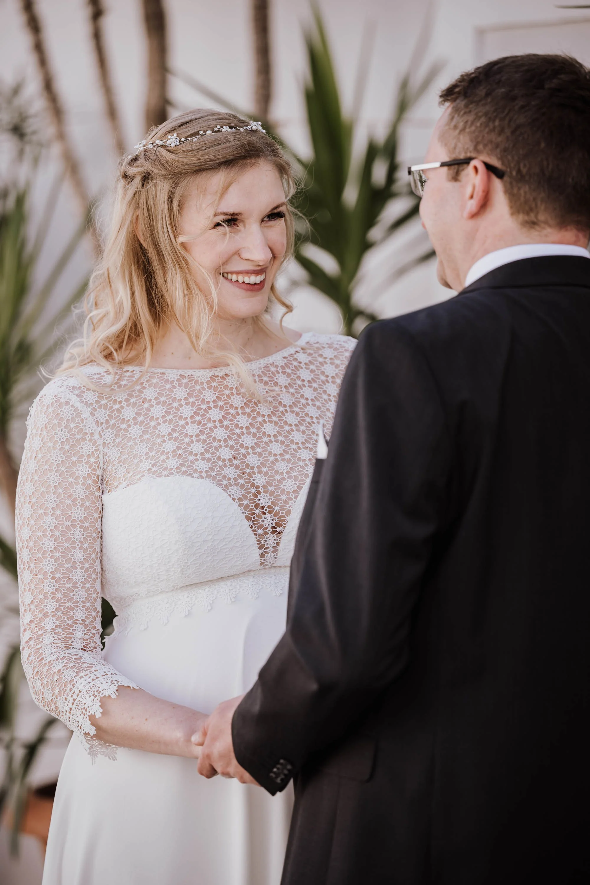 Braut und Bräutigam halten Händchen während ihrer Hochzeit, die Braut trägt ein weißes Kleid mit Spitze und eine Diadem, beide lächeln sich an.