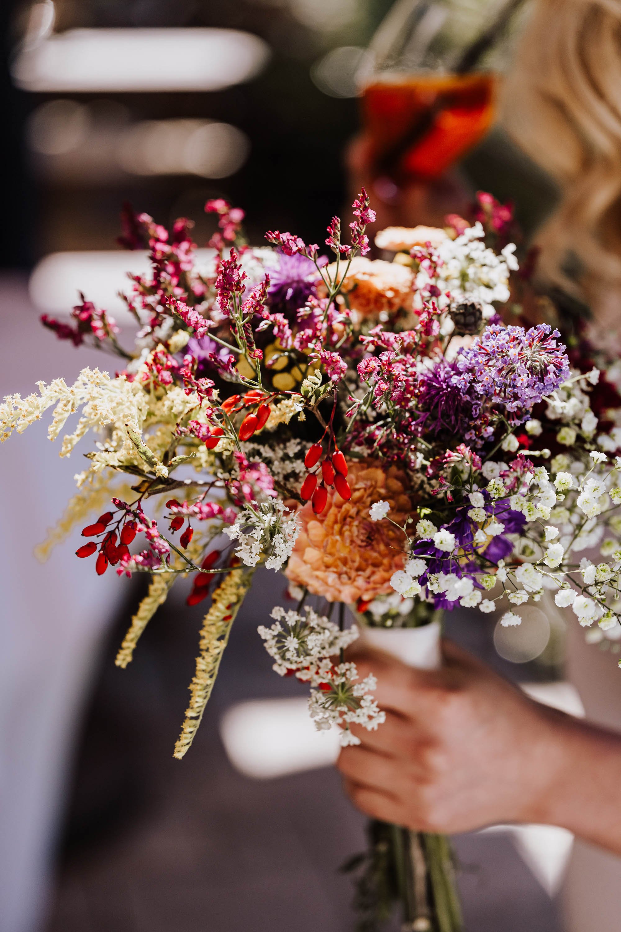 Person hält einen bunten Blumenstrauß mit verschiedenen Blumen und Zweigen. Im Hintergrund ein Glas mit Getränk.