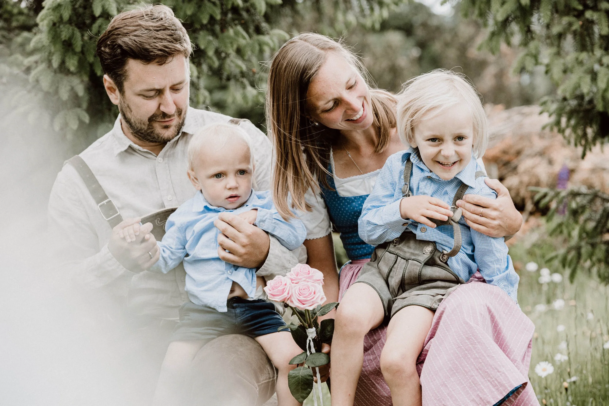 Familie mit zwei kleinen Jungen im Garten, einer hält einen Rosenstrauß, alle lachen und genießen die Zeit zusammen.