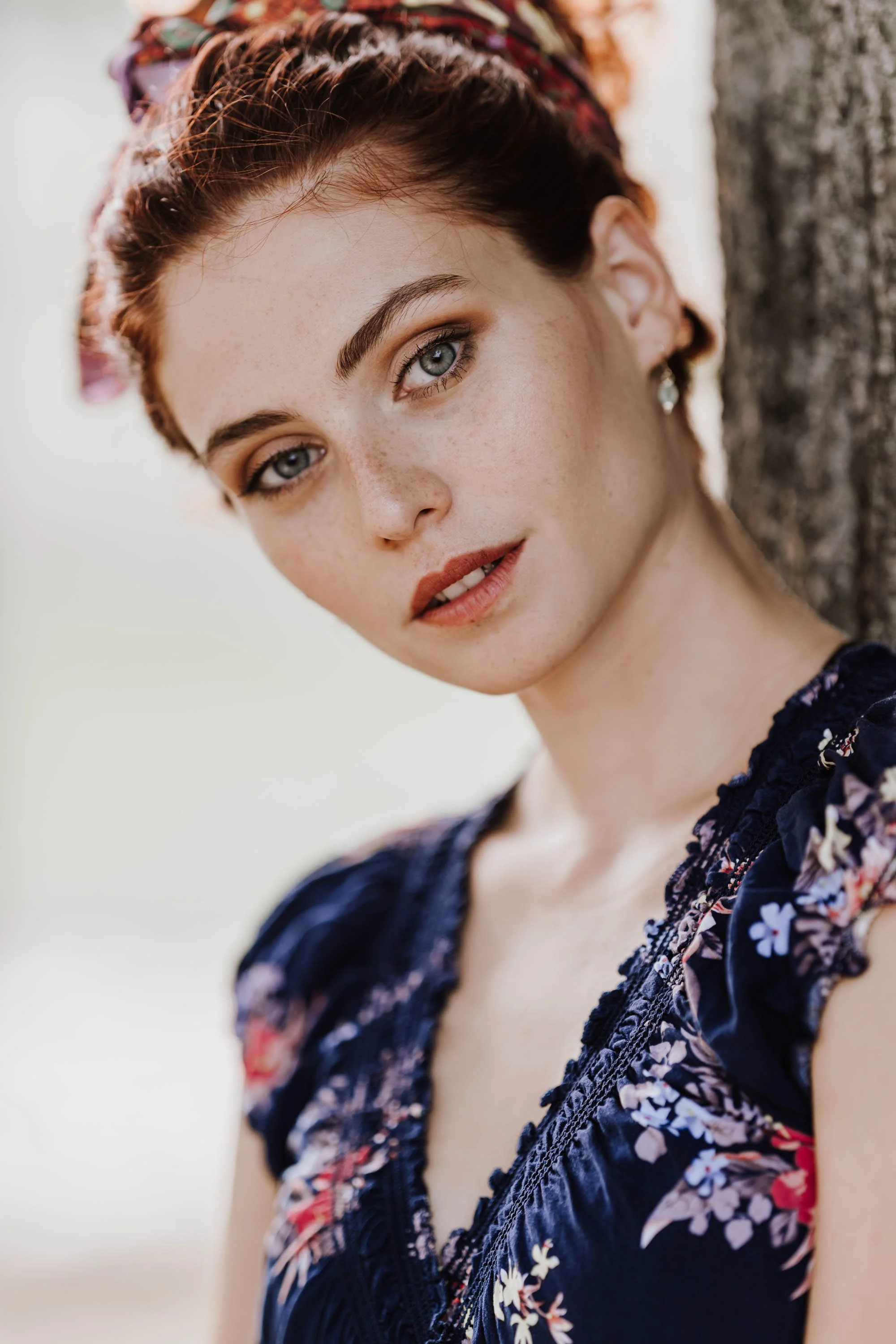 Nahaufnahme eines jungen Mädchens mit blonden, lockigen Haaren, blauen Augen und Sommersprossen, das an einem Baum lehnt, mit einem gedankenvollen Ausdruck.