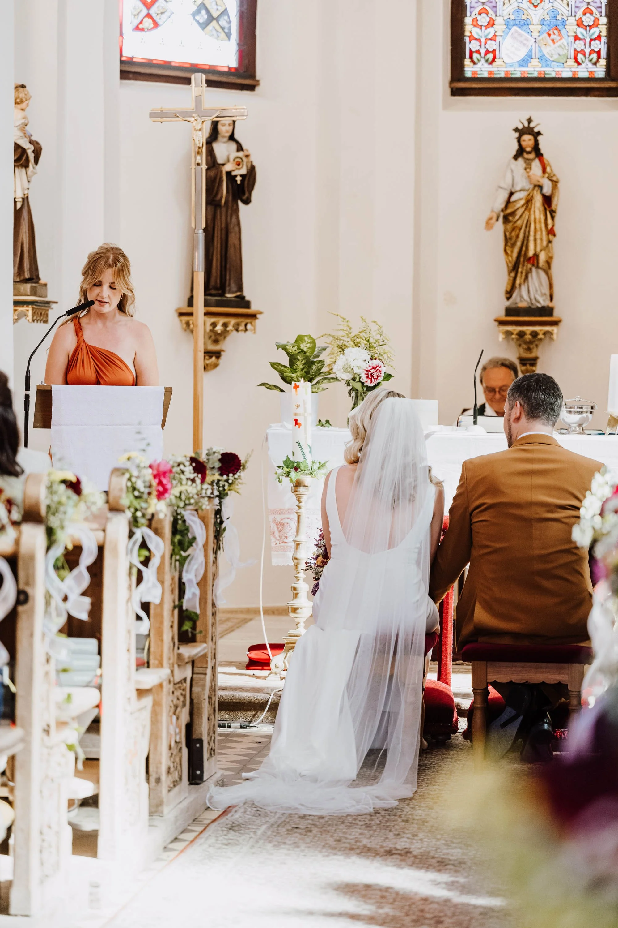 Hochzeit in einer Kirche mit Braut, Bräutigam und einer Frau, die spricht, vor einer Orgel und religiösen Statuen, mit bunten Kirchenfenstern im Hintergrund.