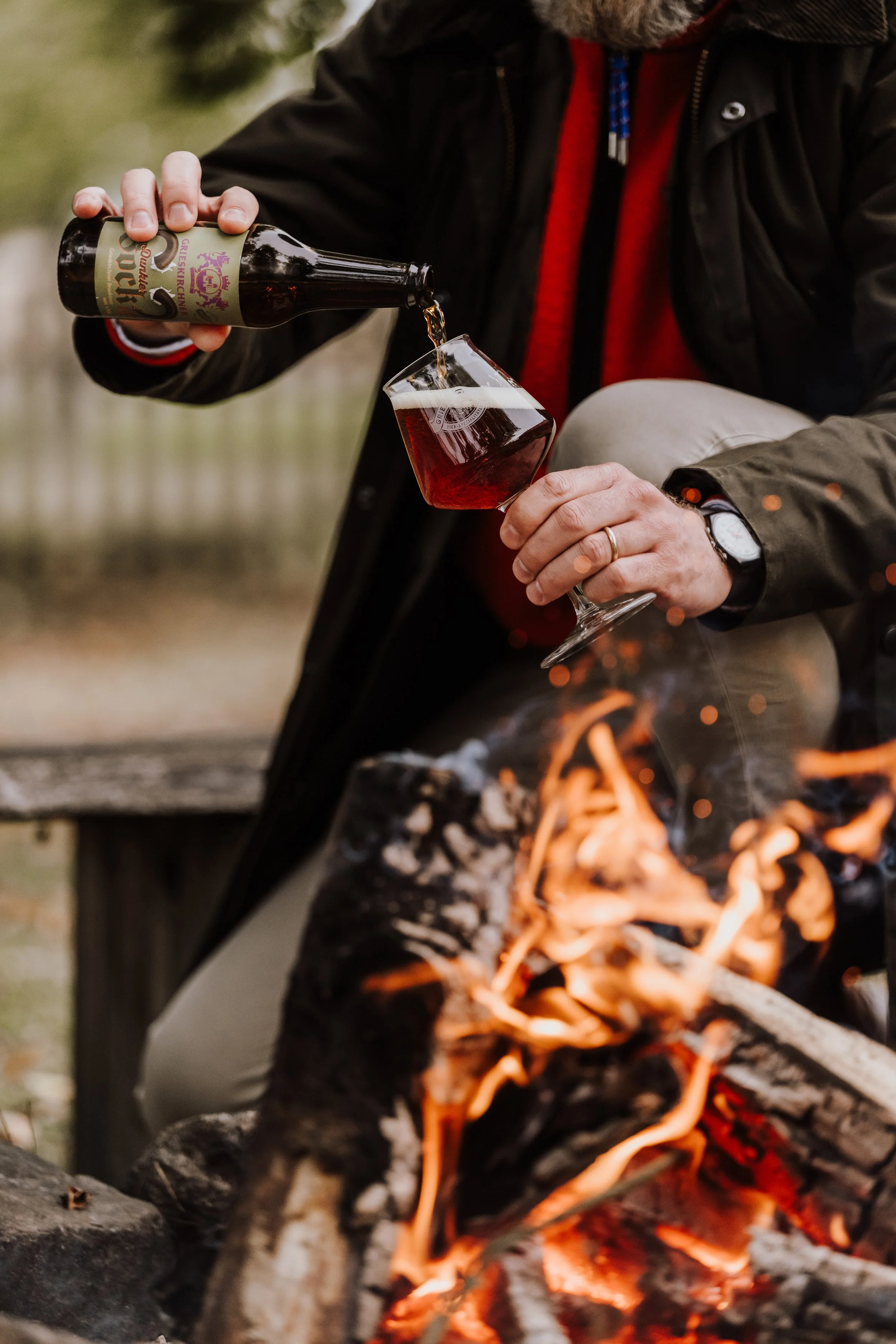 Ein Mensch gießt Wein in ein Weinglas, während er neben einem Lagerfeuer sitzt. Der Mensch trägt eine Jacke, eine Armbanduhr und einen Ring. Die Szene ist im Freien mit einer unscharfen Naturhintergrund.