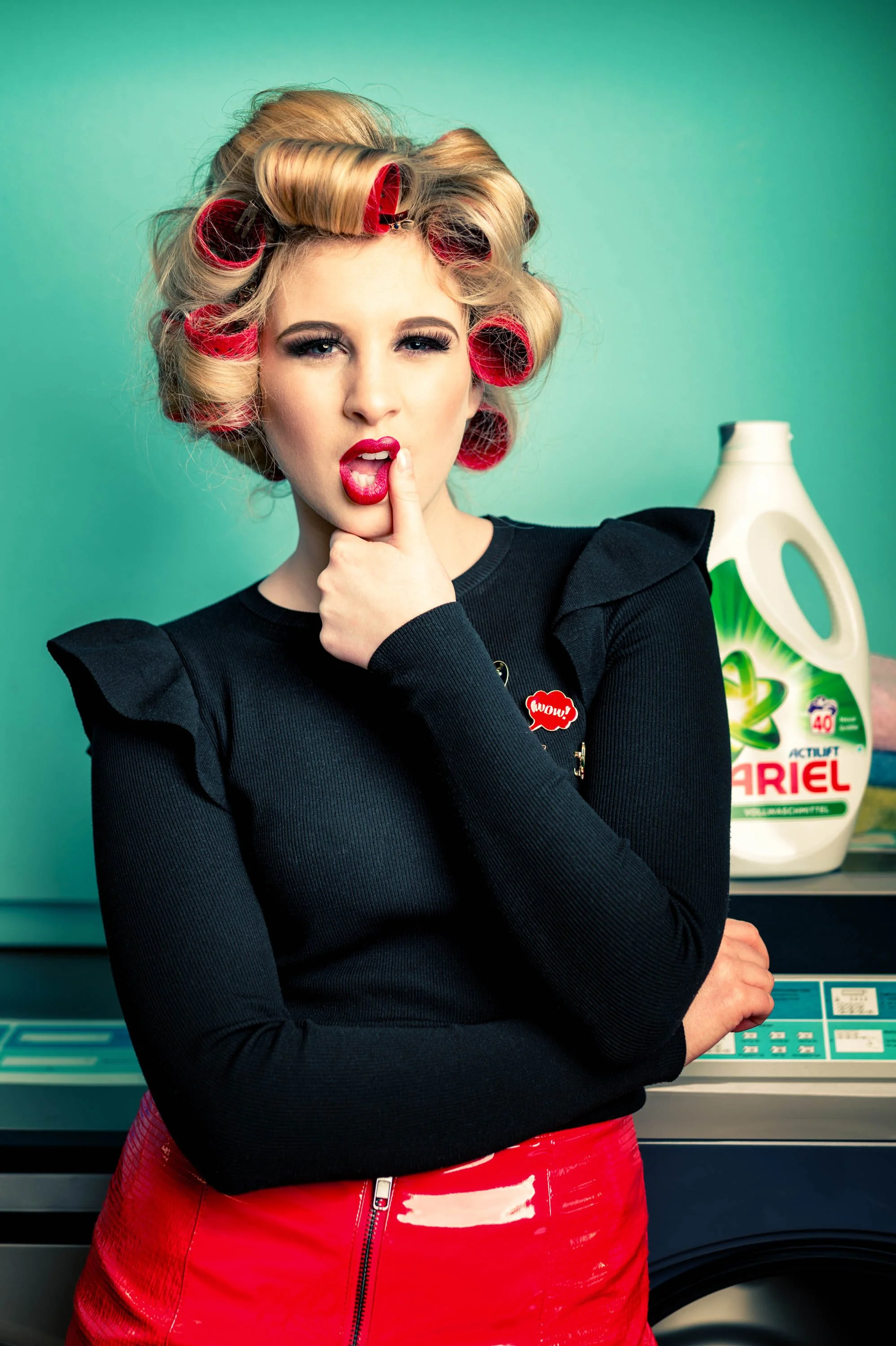 Eine junge Frau mit roten Haarkorken und rotem Lippenstift, die nachdenklich schaut, mit einem Handfinger an die Lippe, vor einem Wäscheautomat und einem Ariel Waschmittel.
