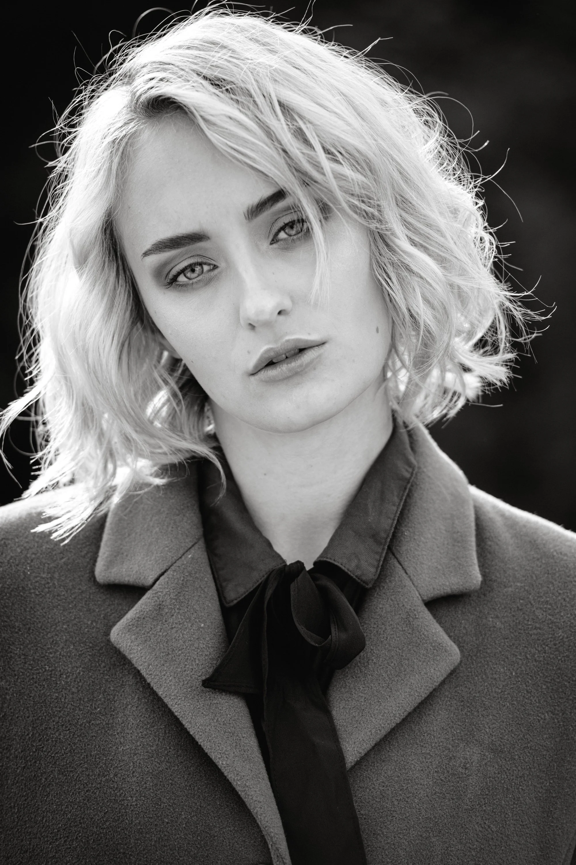 Schwarz-Weiß-Fotografie eines jungen Frau mit lockigem, shoulder-langen Haar, trägt eine dunkle Jacke und eine schwarz gebundene Schleife am Kragen, Blick direkt zur Kamera.