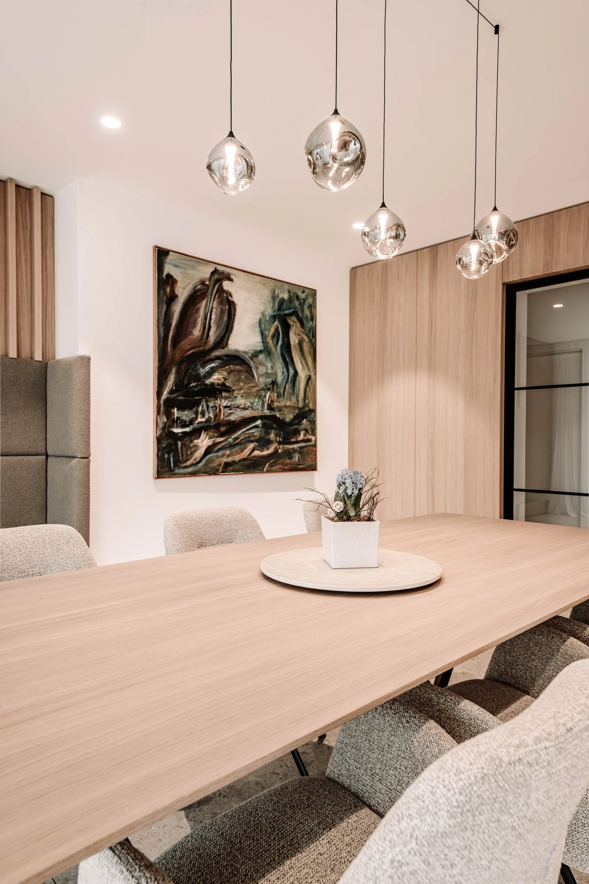 Moderner Esszimmerraum mit einem großen Holz Tisch, mehreren gepolsterten Stühlen, einer Wandkunst, einer dekorativen Pflanze auf einem Teller in der Mitte des Tisches und stilvollen, hängenden Lampelementen.