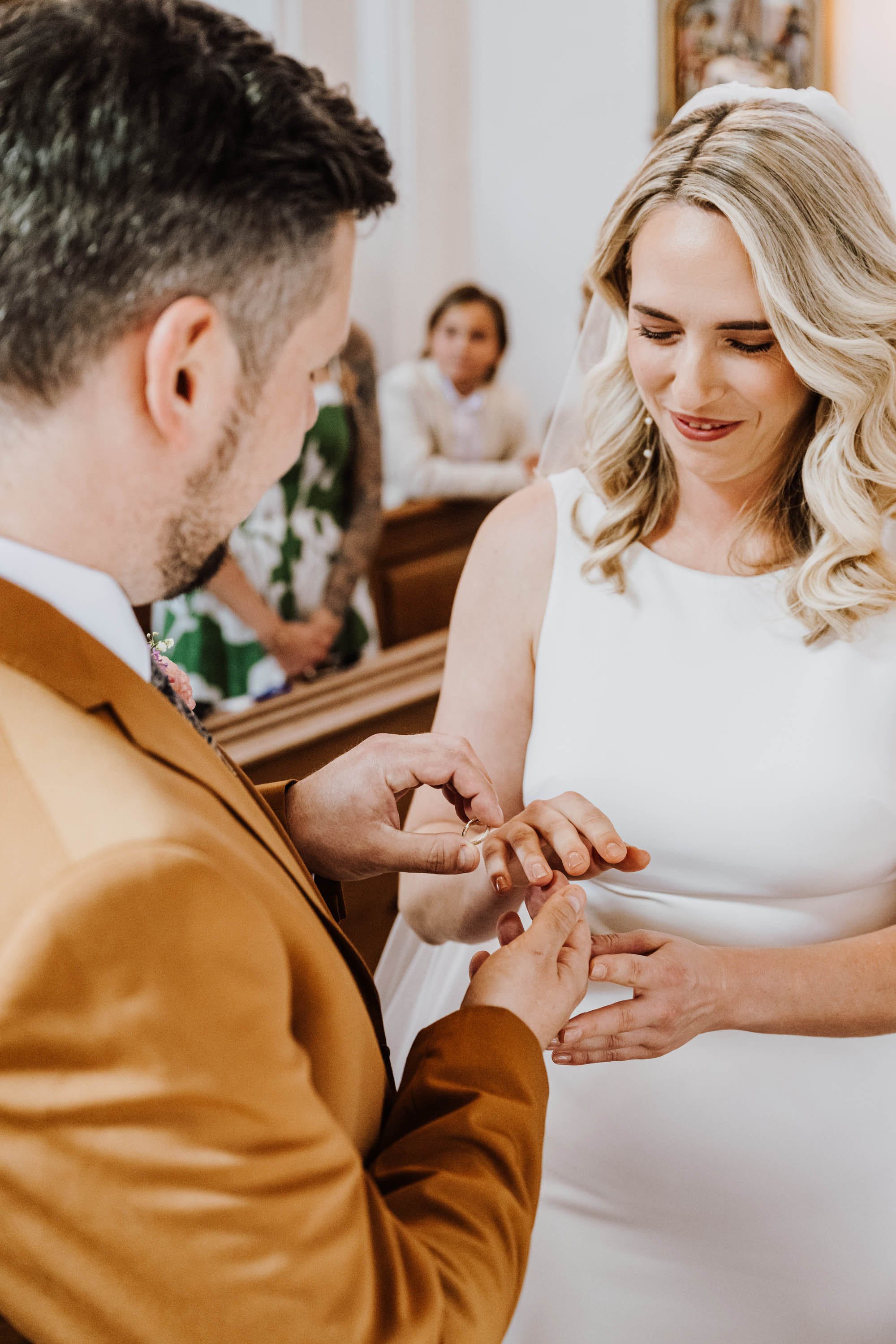 Ein Mann in einem braunen Anzug legt einer Frau einen Ehering an, während sie bei einer Trauung lächelt. Im Hintergrund beobachten weitere Gäste.