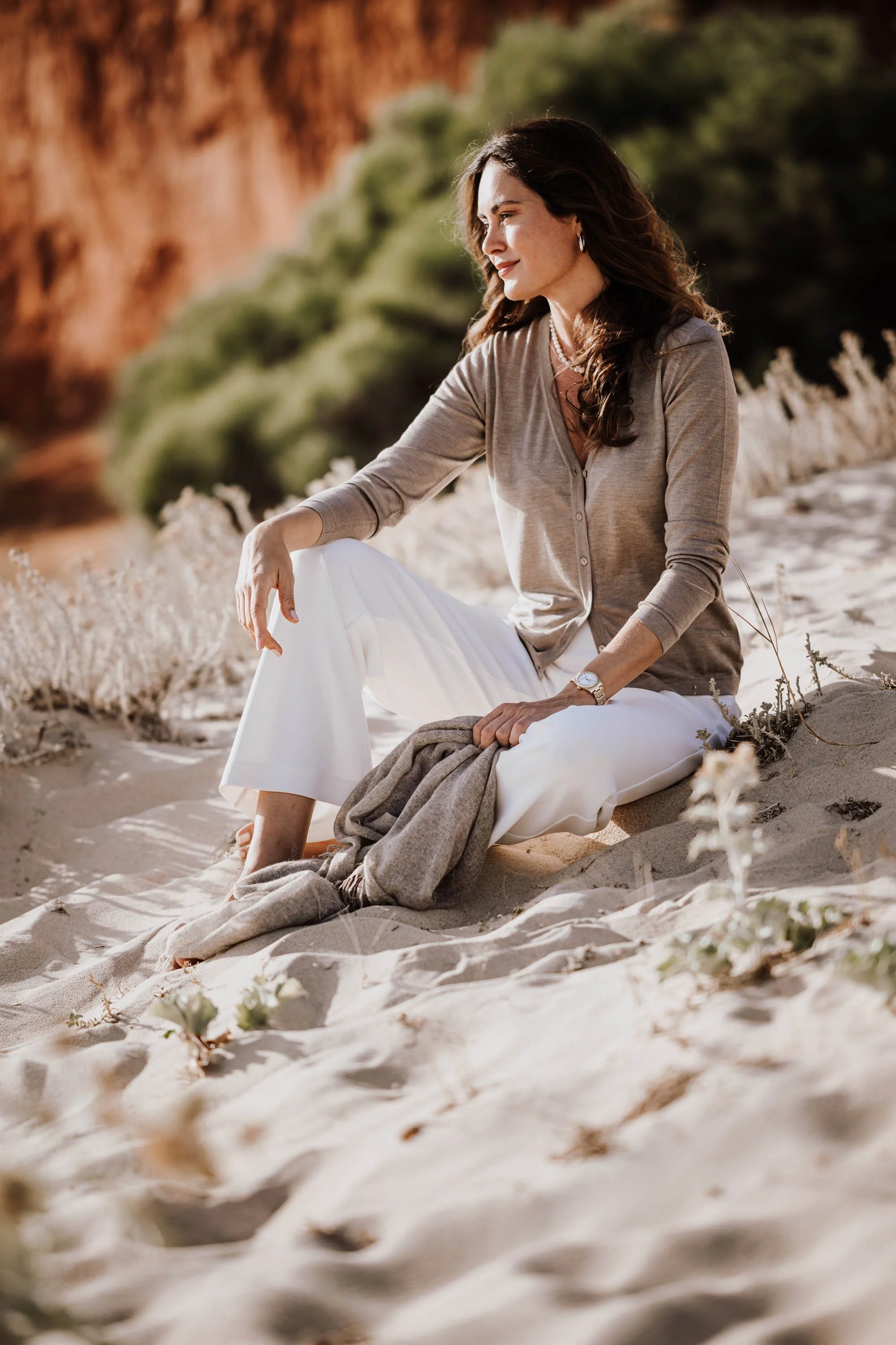 Eine Frau sitzt entspannt am Strand im Sand mit einer Landschaft aus grünen Büschen und roten Felsen im Hintergrund. Sie trägt eine beige Bluse, weiße Hosen und eine Uhr, wirkt nachdenklich und genießt die Natur.
