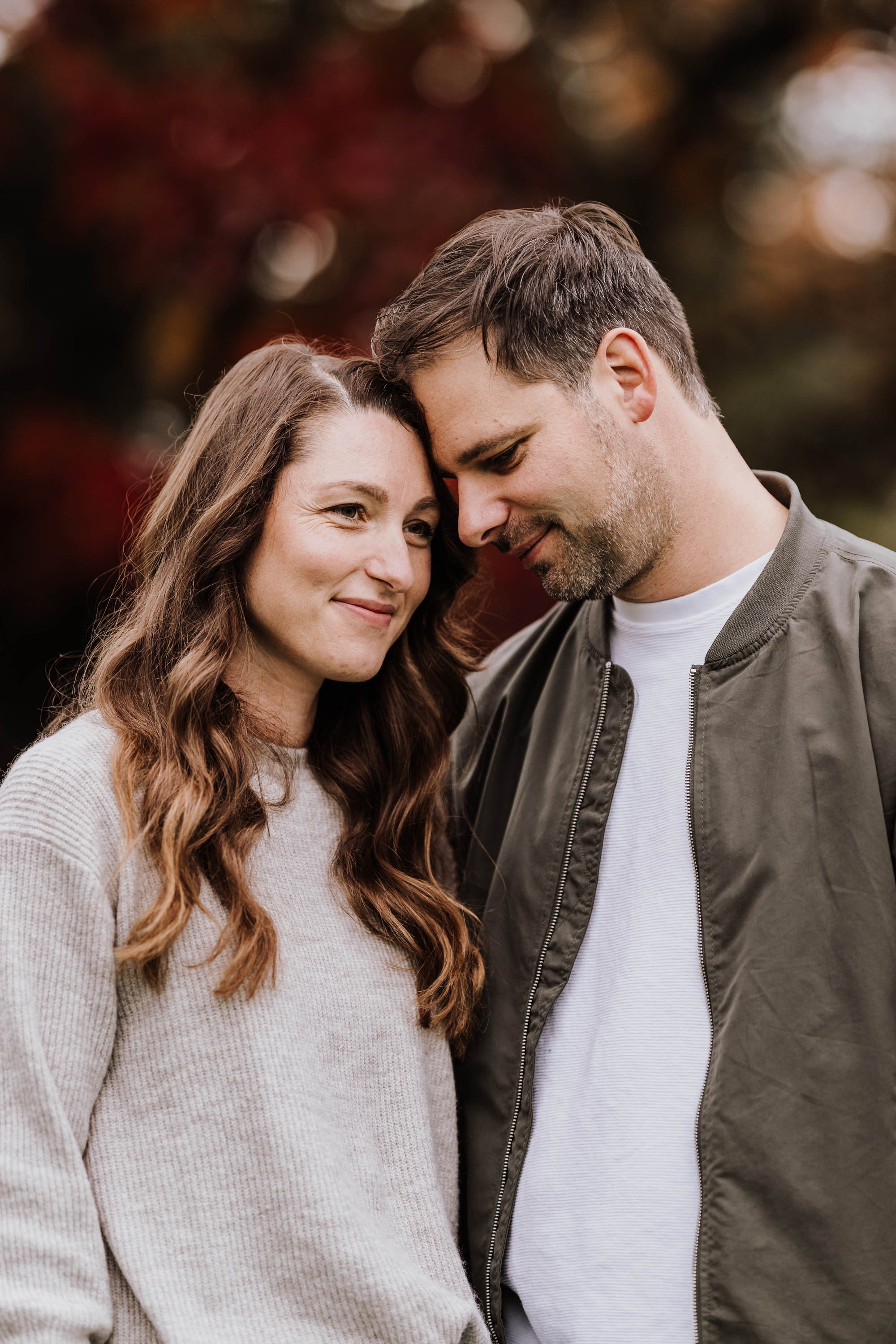 Ein Paar steht eng beieinander, lächelt und hat die Stirn aneinander, im Hintergrund bunte Herbstblätter.