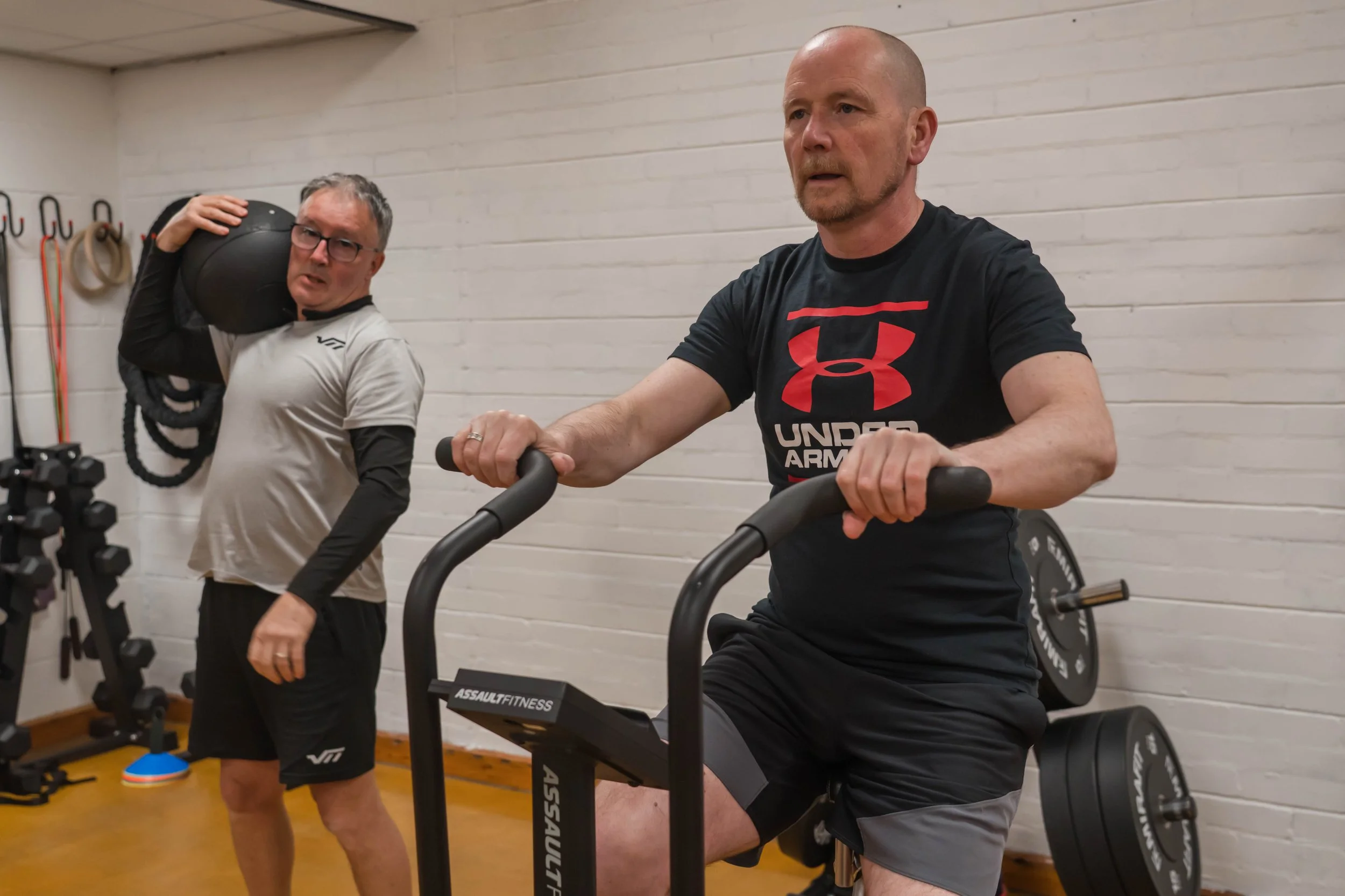 Two men are working out at Infinity Coaching . One man, wearing a black Under Armour T-shirt and gray shorts, is on a stationary bike. The other man, wearing glasses and a light-colored shirt, is holding a black medicine ball over his shoulder.