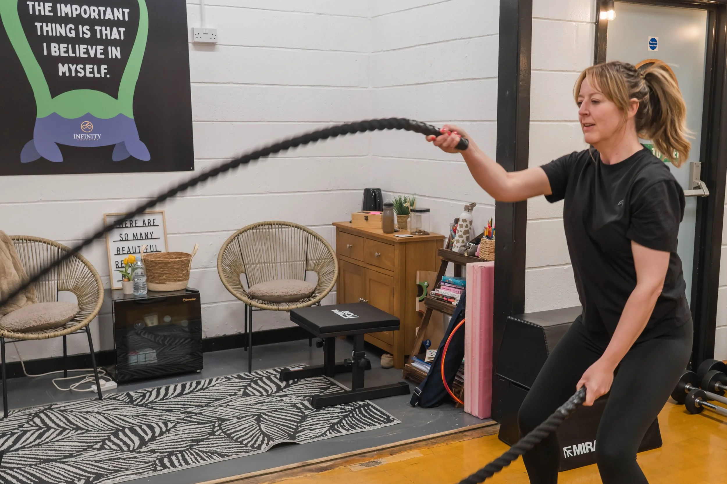 A woman with blonde hair in a ponytail is exercising with battle ropes inside a room decorated with wicker chairs, plants, and framed posters. She is wearing a black T-shirt and black leggings, and is holding one end of a thick, black rope, swinging it in a workout movement.