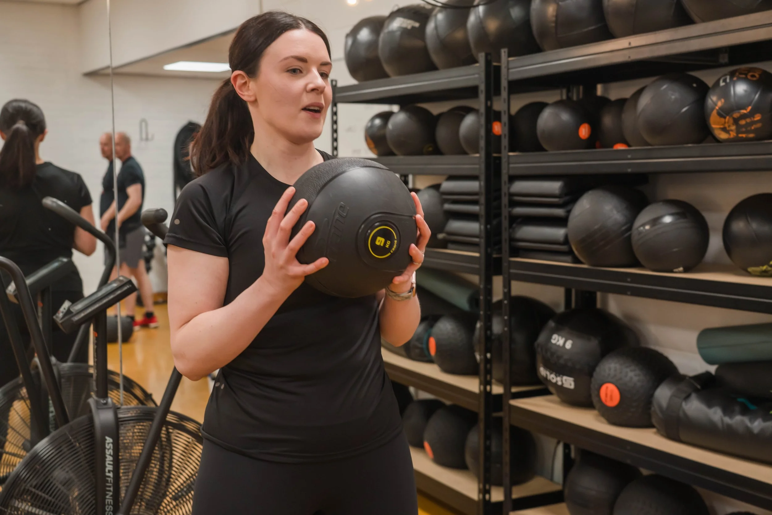 A woman in black workout clothes holding a black medicine ball in a gym with shelves of exercise balls and mats.