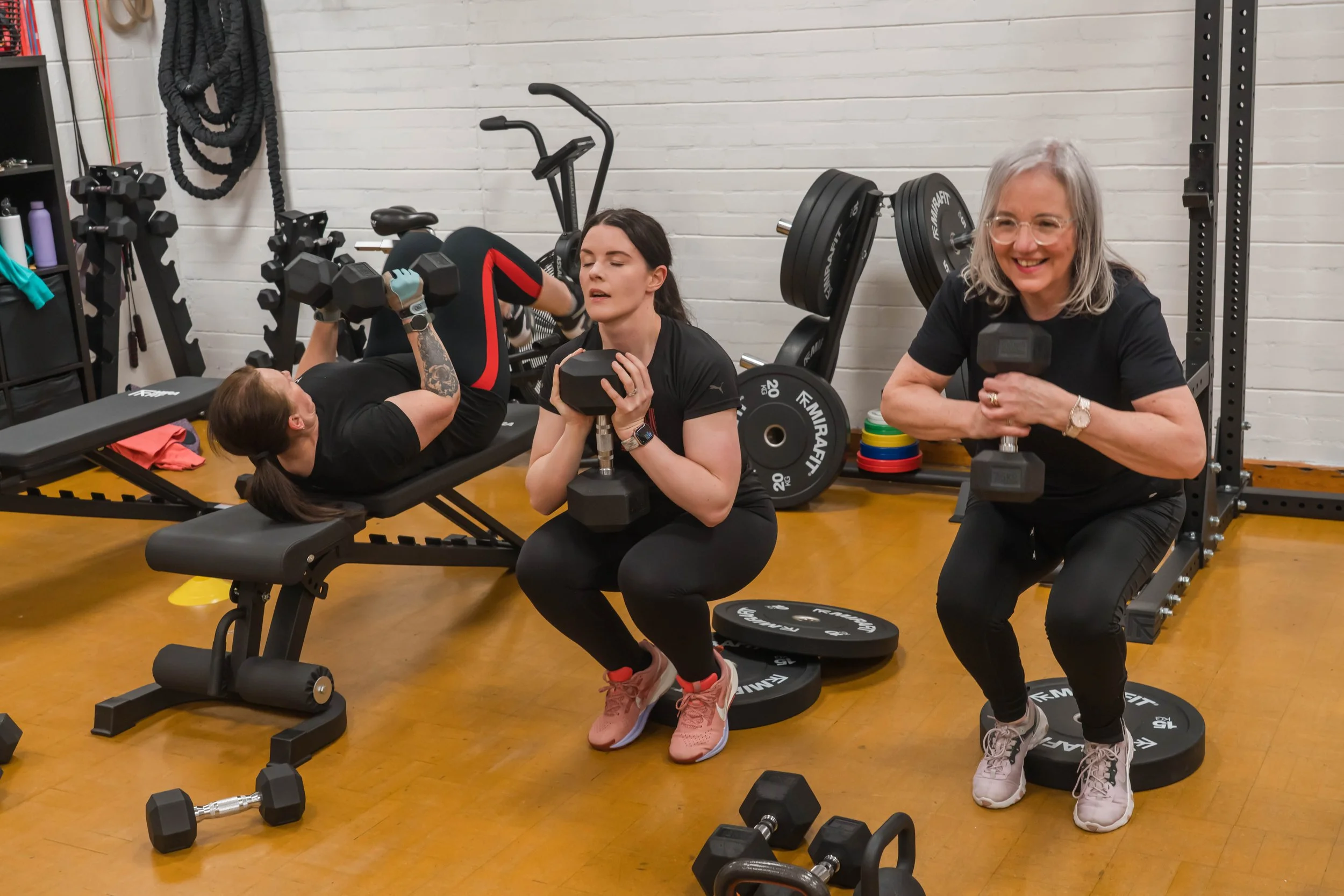 Three women working out with dumbbells in a gym. one woman is lying on a bench, lifting dumbbells, while the other woman is standing, holding a dumbbell and smiling. The third woman is kneeling on the ground, holding a dumbbell close to her chest, with her eyes closed.