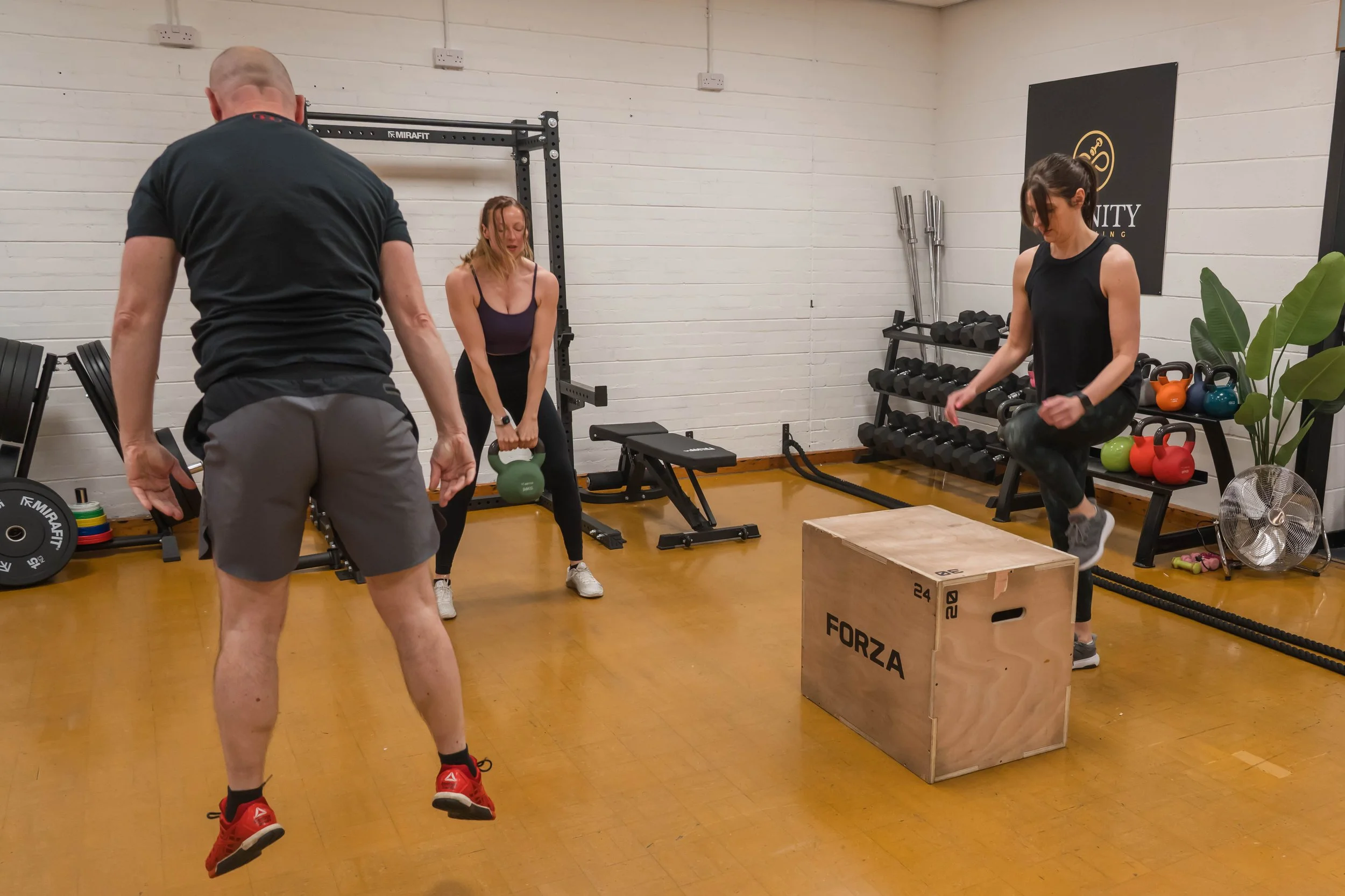 A group workout class in a gym with two women holding kettlebells and a man instructing. One woman is jumping onto a wooden plyometric box, while the other woman looks down, possibly preparing to lift the kettlebell. The gym walls are lined with weights and fitness equipment.