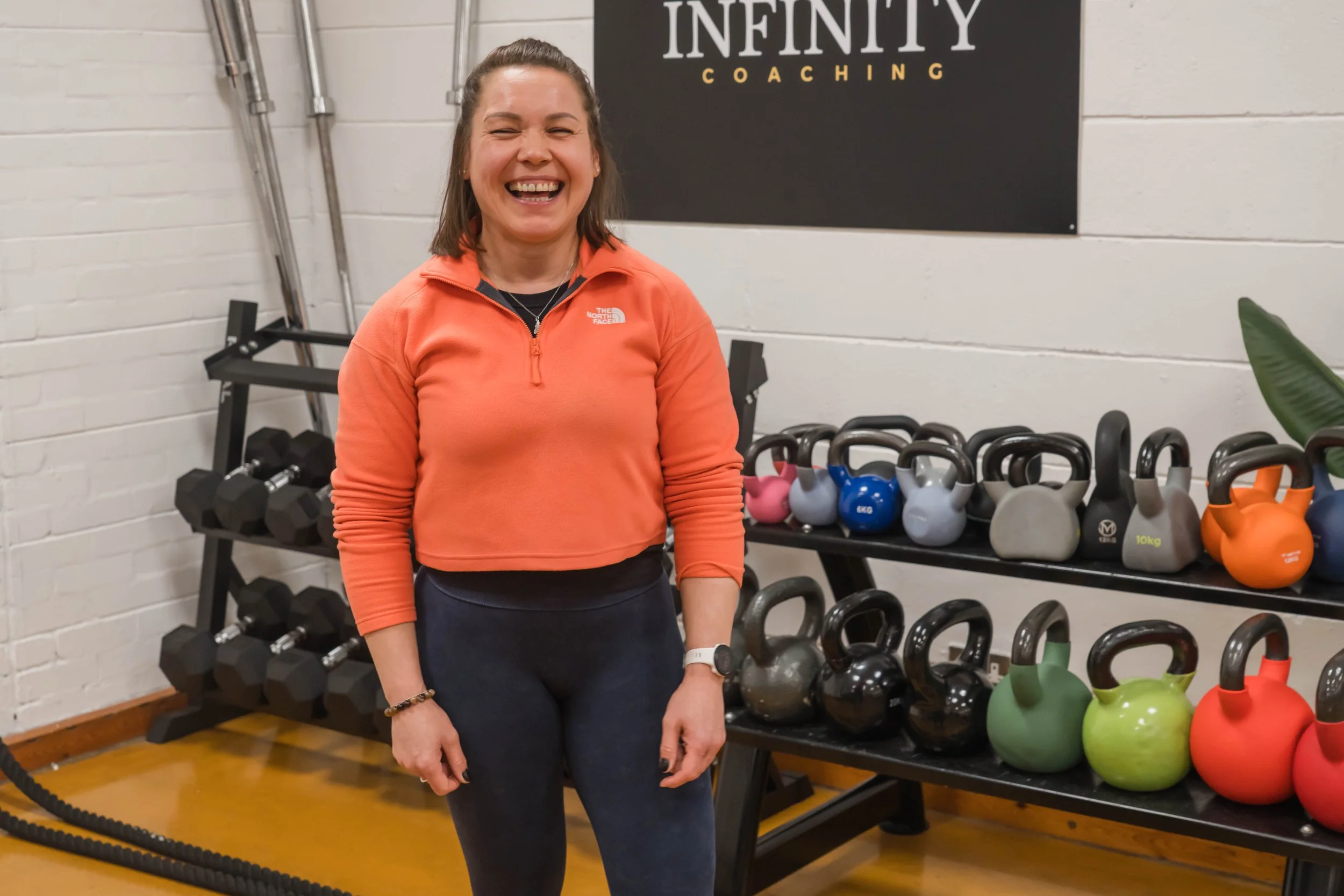A smiling woman in an orange jacket standing in a gym with black and colorful kettlebells and dumbbells on racks behind her. A sign reading 'Infinity Coaching' is visible on the wall.