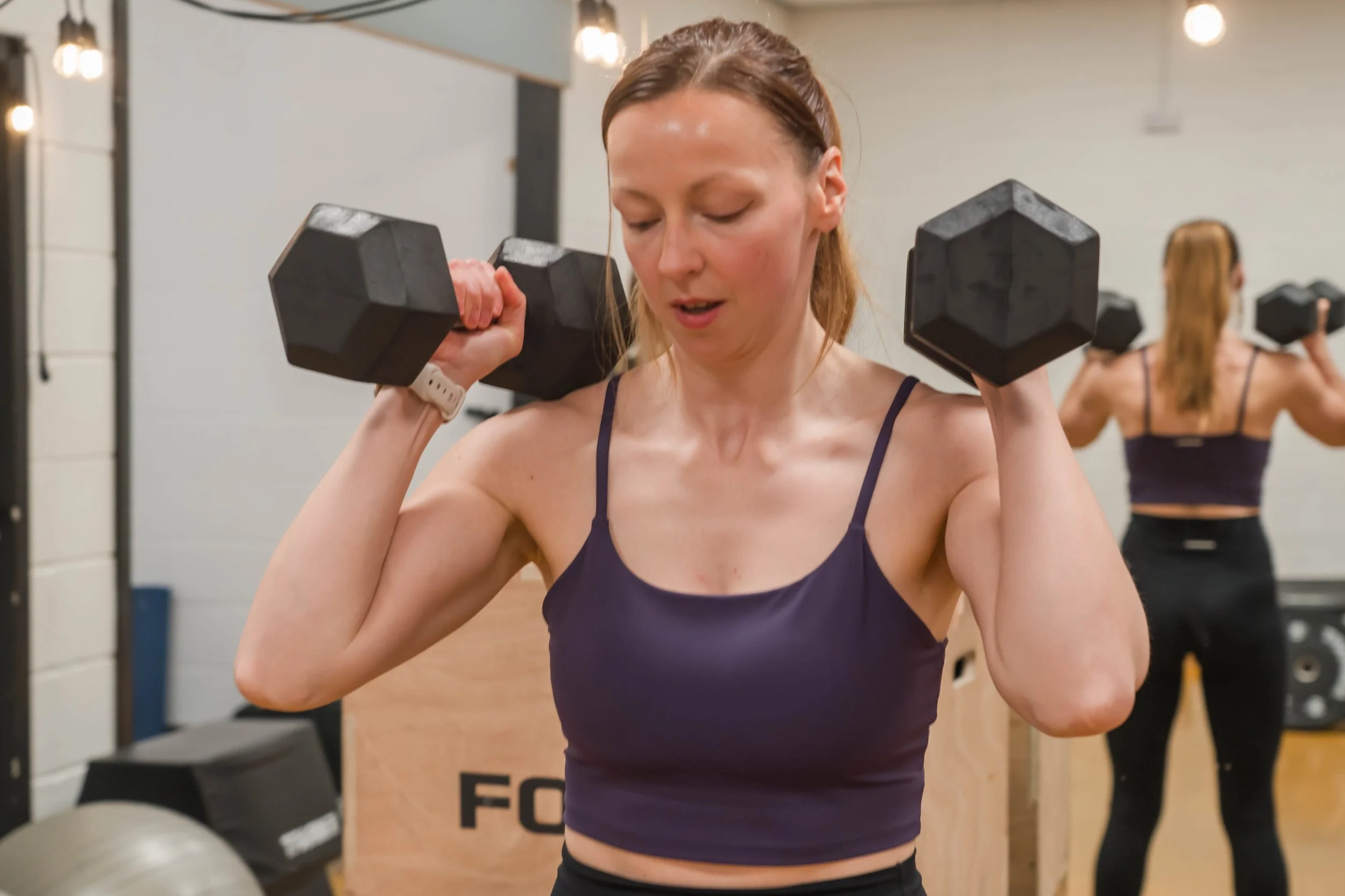 A woman with light skin and long, reddish-brown hair tied back, wearing a navy tank top, is lifting black dumbbells in a gym. She has a focused expression and is looking slightly downward. In the background, another woman is also lifting dumbbells, and gym equipment such as a rubber stability ball and weight plates are visible.