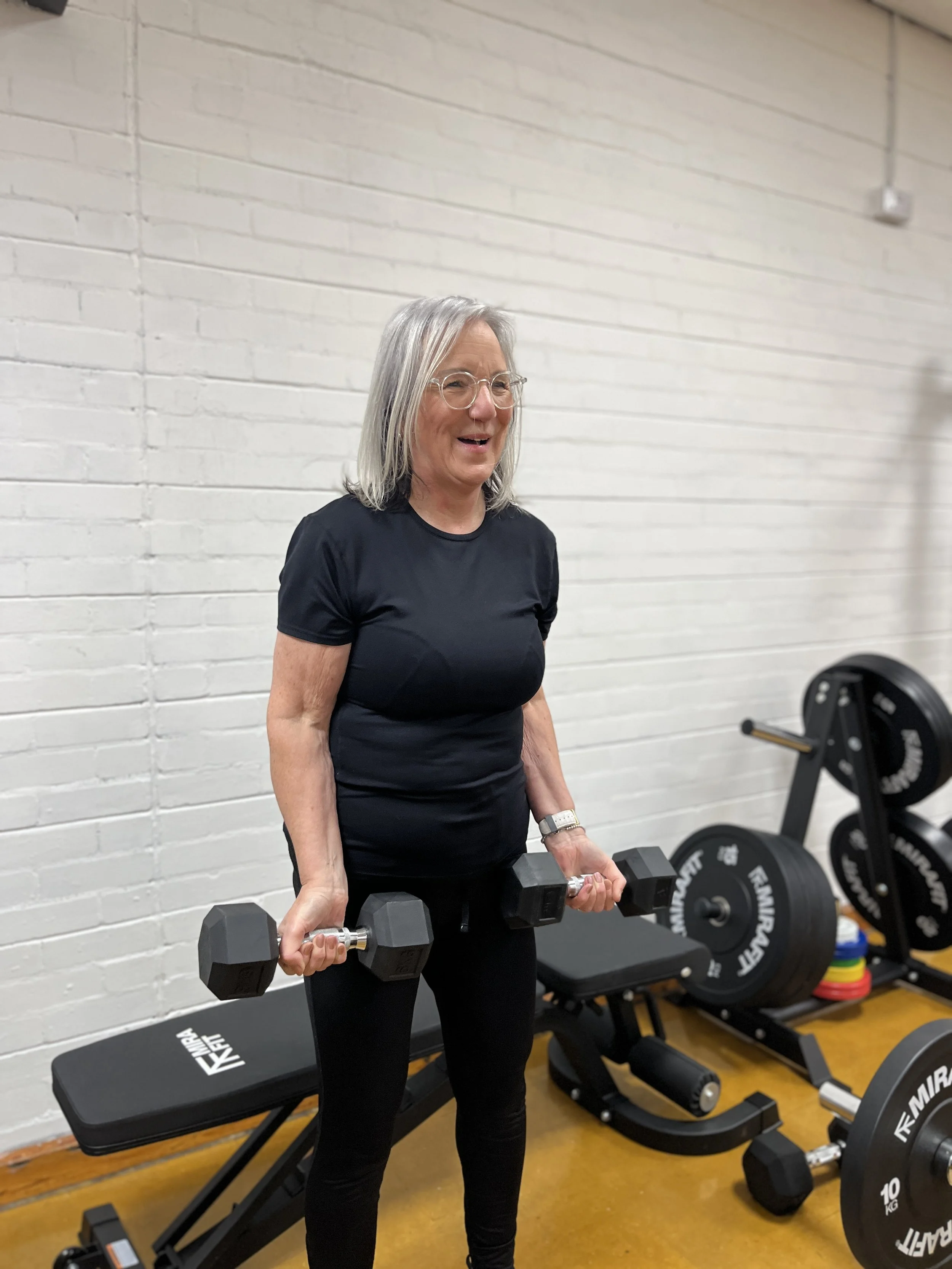 An older woman with grey hair, glasses, wearing a black shirt and pants, is holding two black dumbbells while standing in a gym. There are weightlifting equipment and plates in the background, including a seated bench and weight plates on a rack.