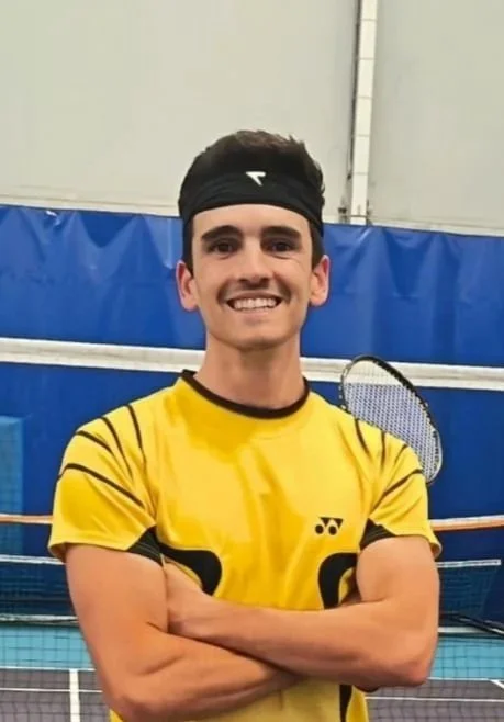 Jeune homme souriant portant un bandeau noir et un t-shirt jaune de sport, tenant une raquette de badminton, sur un terrain intérieur de badminton.