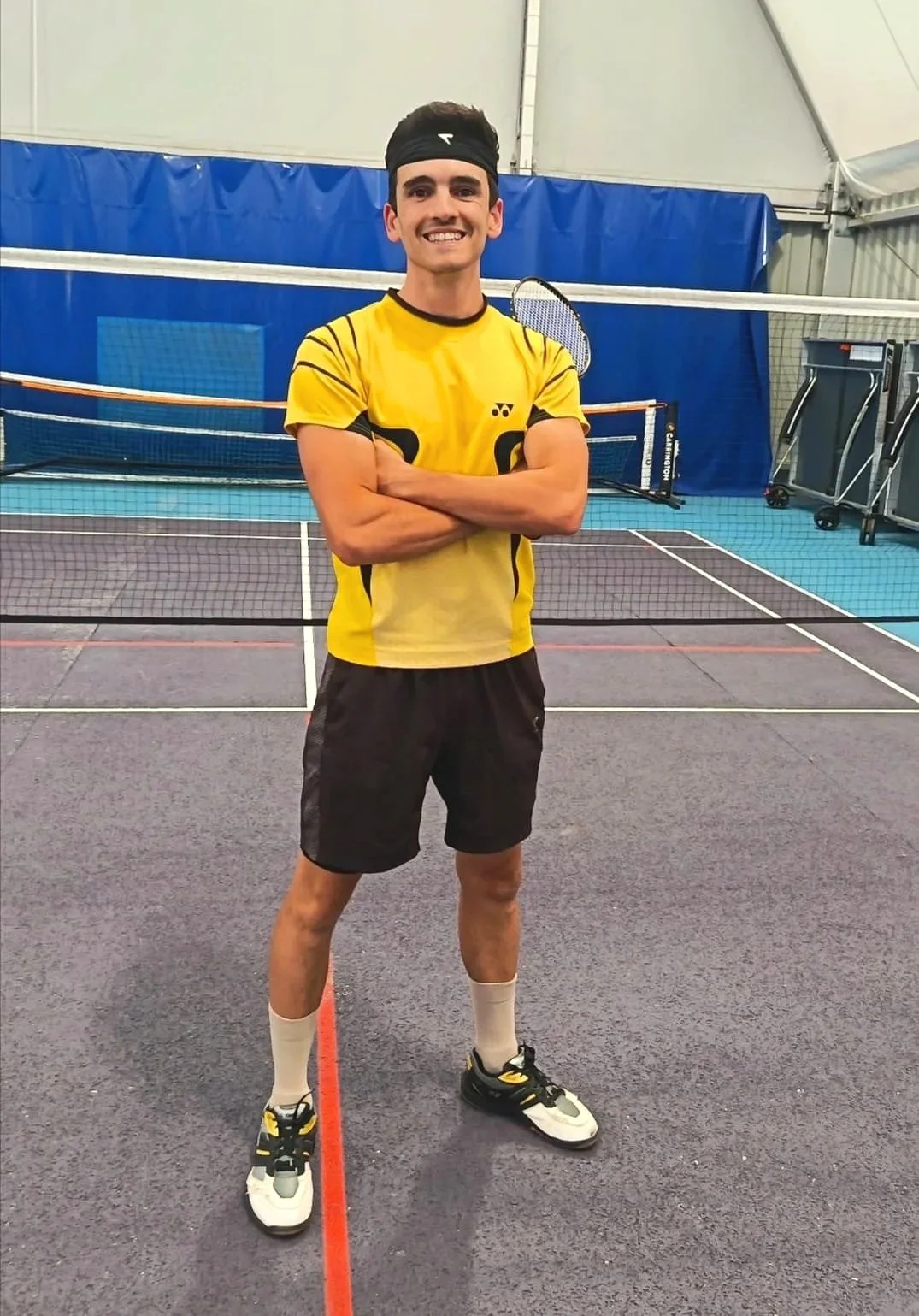 Un jeune homme souriant portant un maillot de sport jaune, un short noir, des chaussures de tennis, des chaussettes blanches, et un bandeau noir, se tient sur un terrain de tennis en intérieur, avec des raquettes et un filet en arrière-plan.