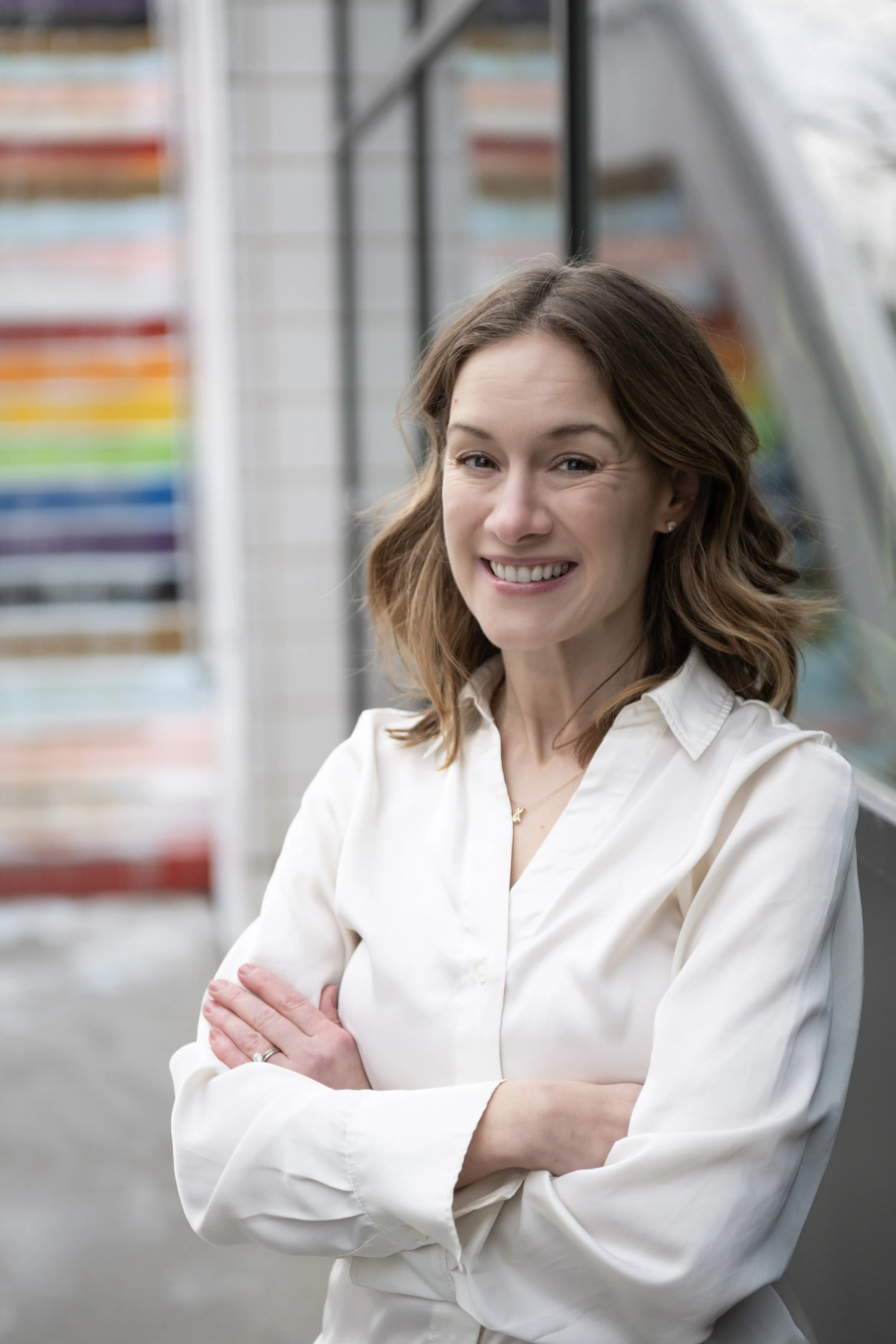 A woman with shoulder-length brown hair, wearing a white button-up shirt, smiling with her arms crossed, standing outdoors in front of a rainbow-colored wall.