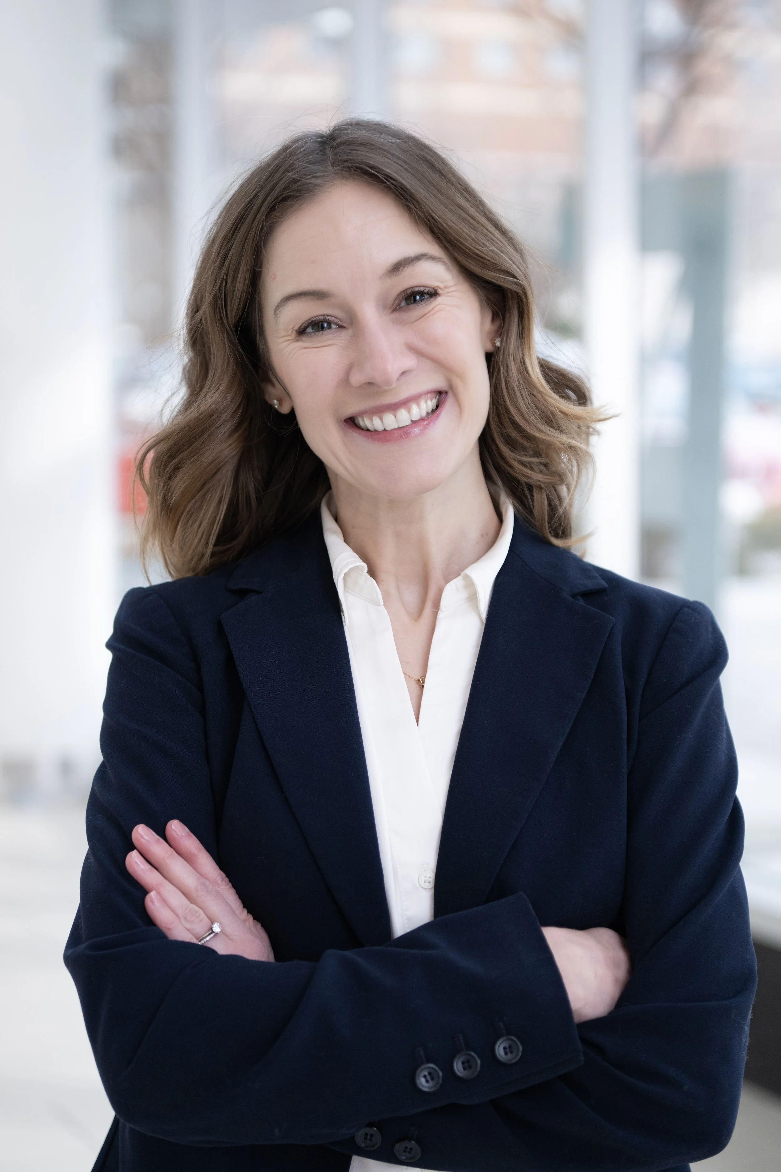 A woman smiling with her arms crossed, wearing a navy blazer and white shirt, standing indoors against a blurred background of windows and buildings.
