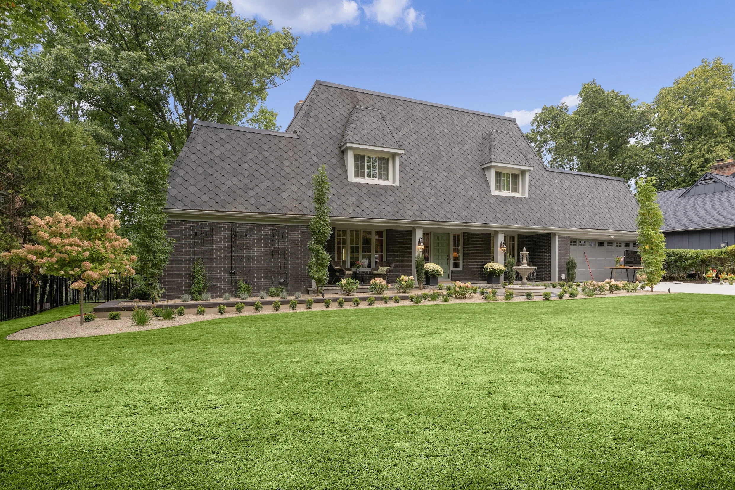 A large house with a gray, shingled roof, dark brick exterior, and green front door. The backyard has a lush, well-maintained lawn, a garden with small plants and trees, and a stone fountain. There are outdoor seating areas and surrounding trees.