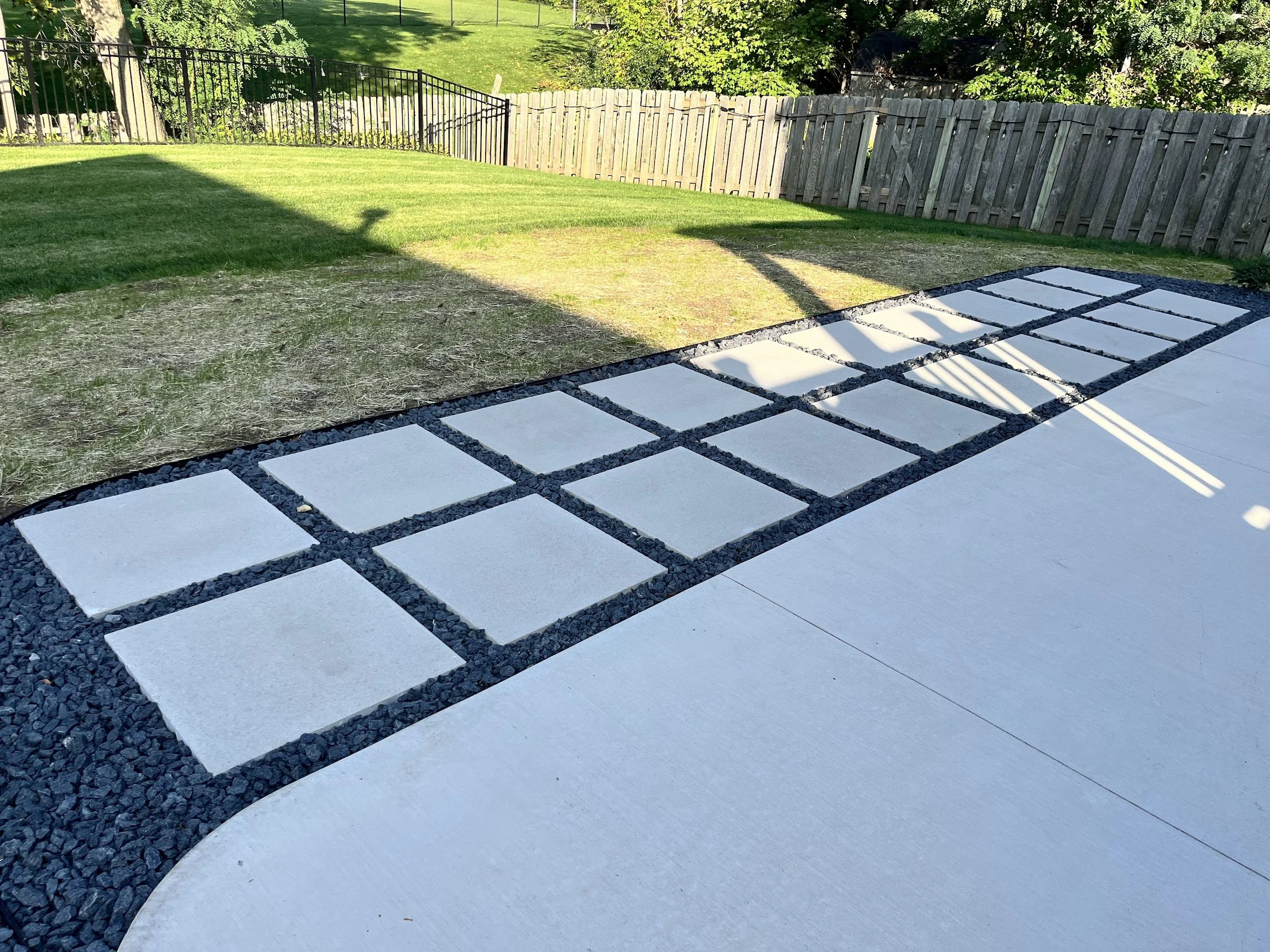 View of a backyard with a patio area featuring white square tiles set in black gravel, grass lawn, and a wooden fence illuminated by sunlight.