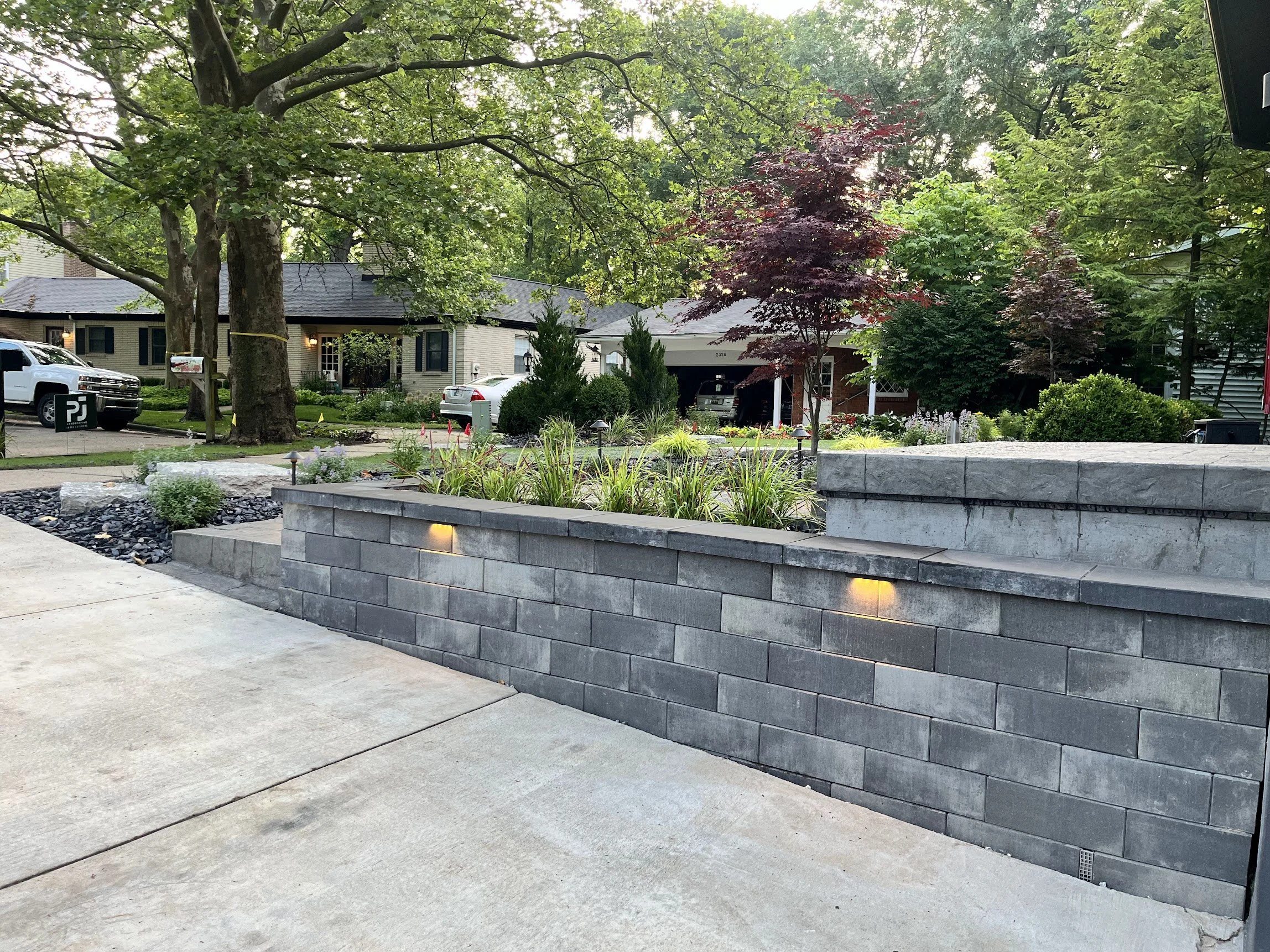Residential backyard with a stone retaining wall, landscaped with plants and decorative lighting, a tree, and neighboring houses visible in the background.