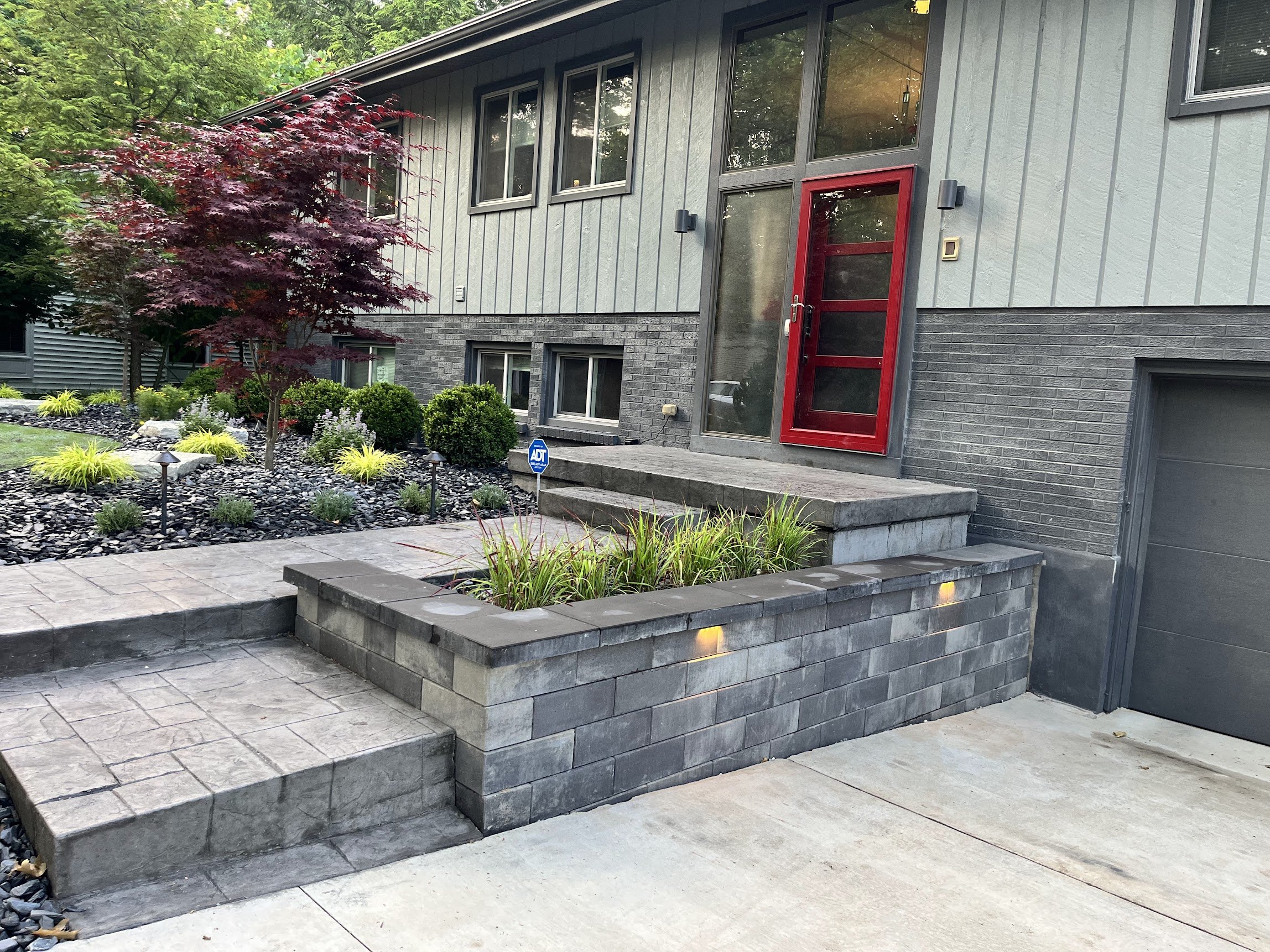 front yard of a modern house with a red door, grey siding and brick walls, landscaped with various green plants, small trees, rocks, and lights.