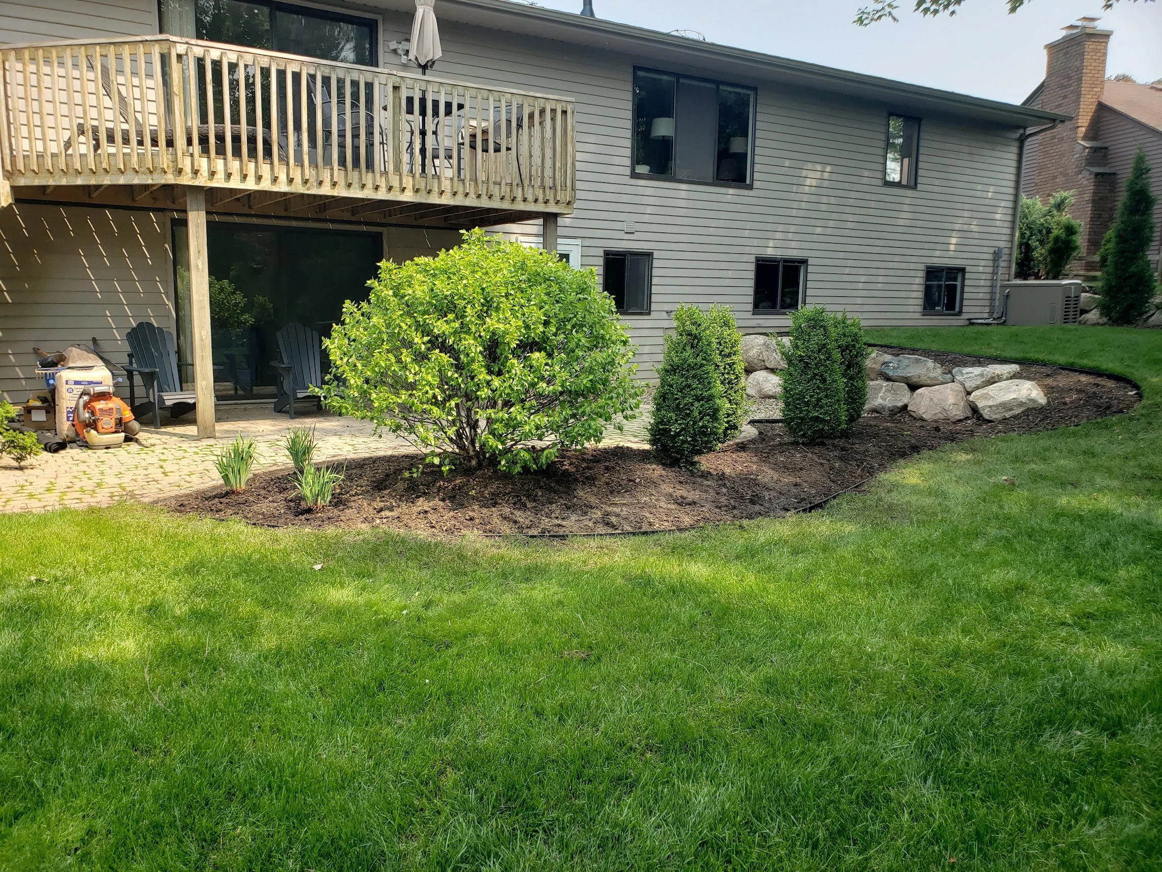 Backyard with green grass, a landscaped area with plants and rocks, and a two-story house with a balcony and patio furniture.
