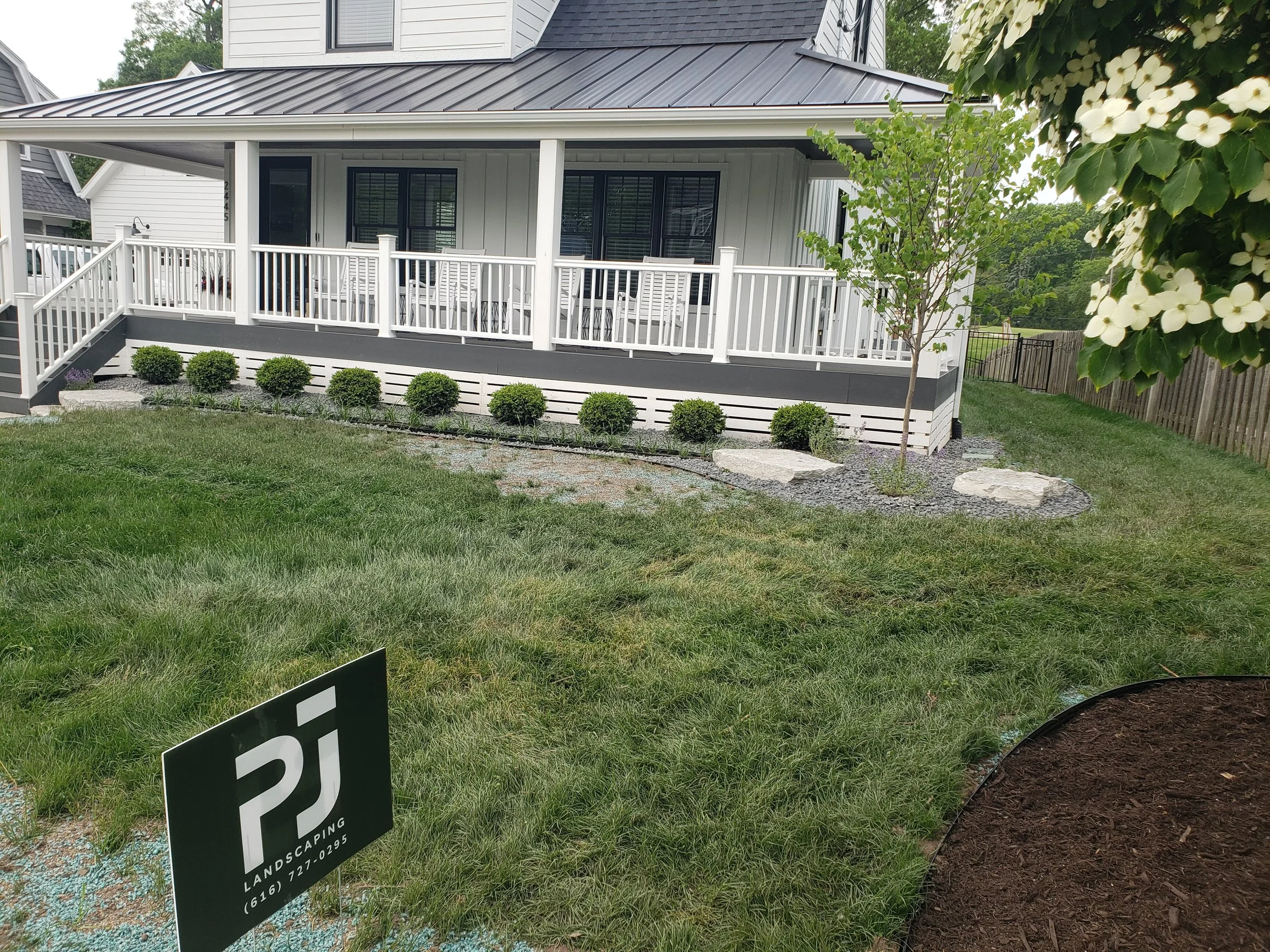 Well-maintained backyard with green grass, a small tree, and a garden bed with mulch. A white house with a raised deck with white railings, a row of neatly trimmed bushes, and a sign that reads 'RJ Landscaping' with a phone number is visible in the p