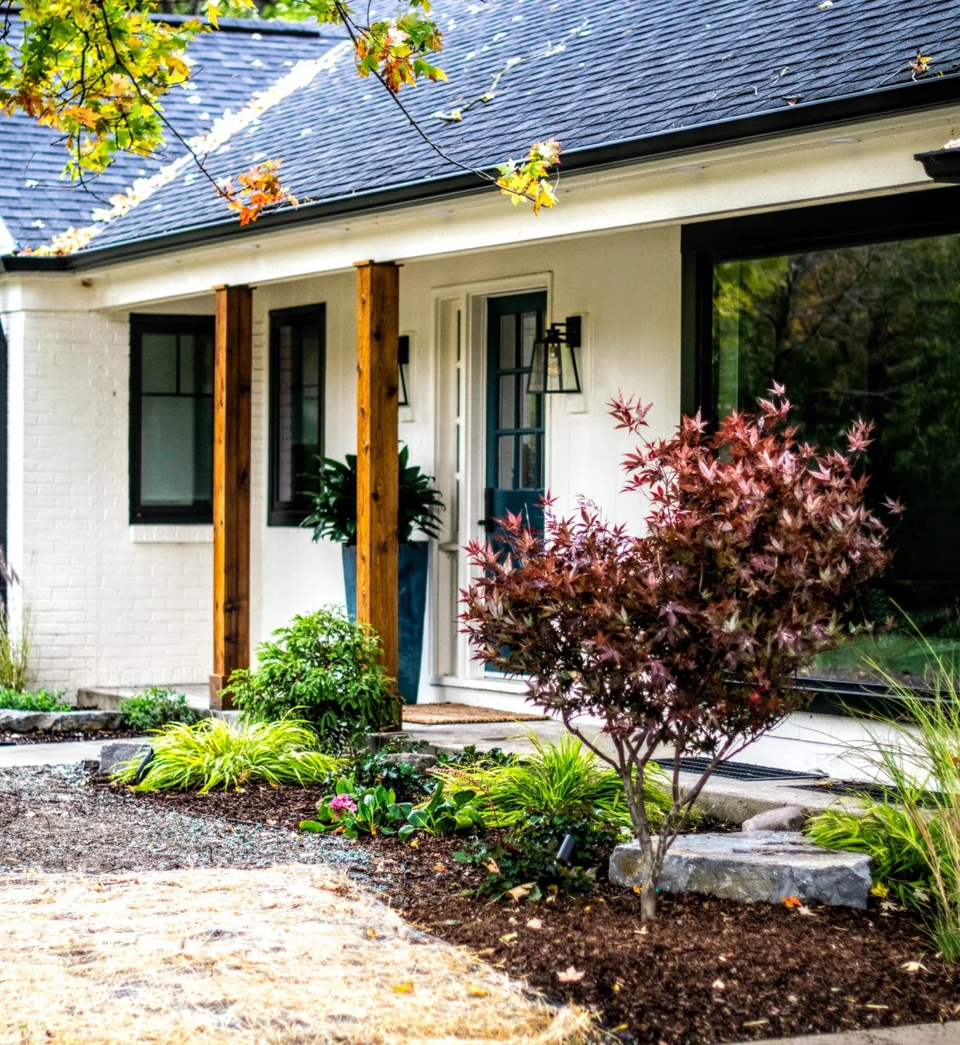 Front yard of a house with a garden, shrubs, and a small tree near the entrance, featuring a white exterior, black window frames, and a wooden post.