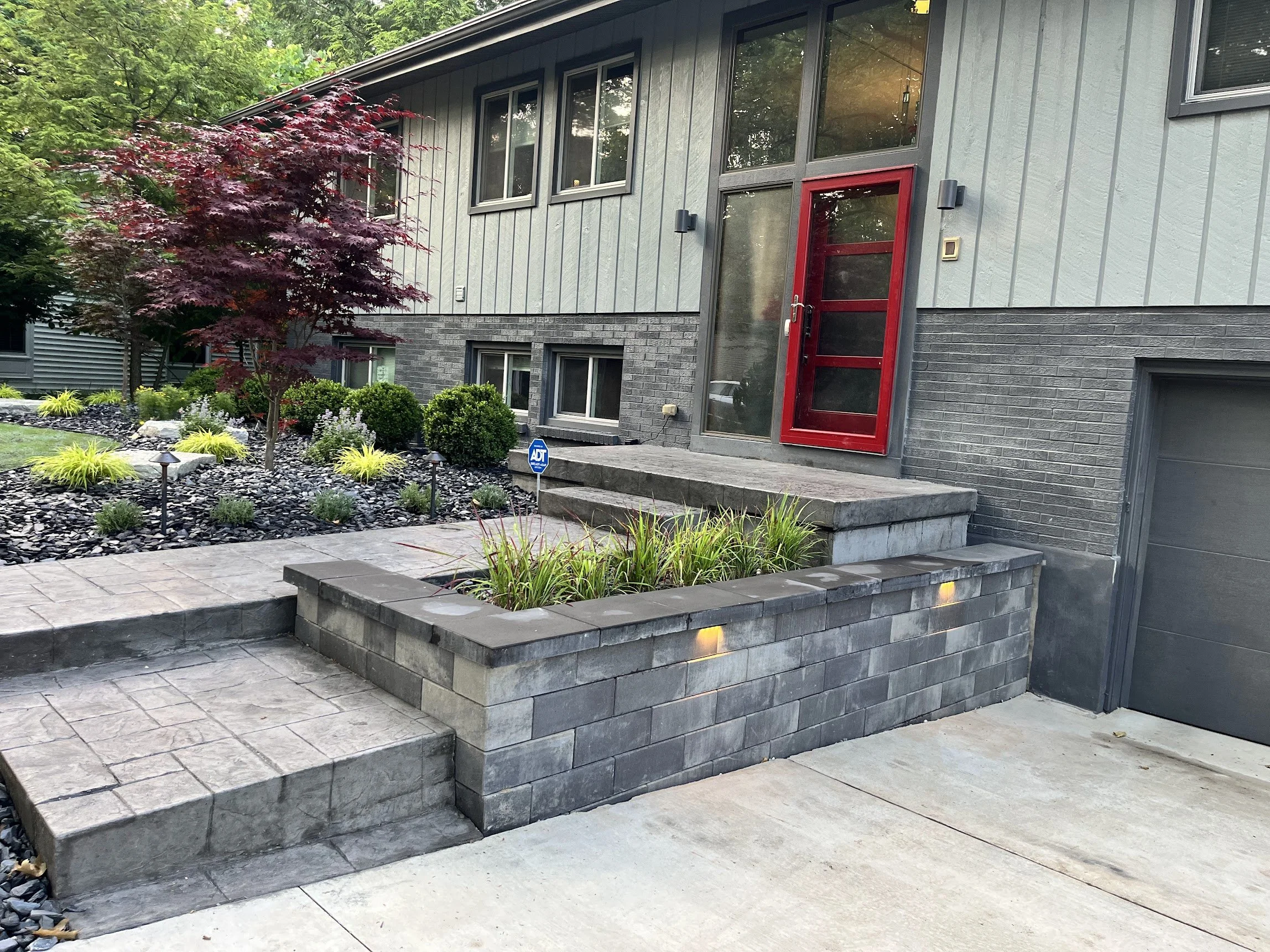 Modern house with gray siding, brick accents, and a red front door, surrounded by landscaped garden with shrubs, a tree, and outdoor lighting.