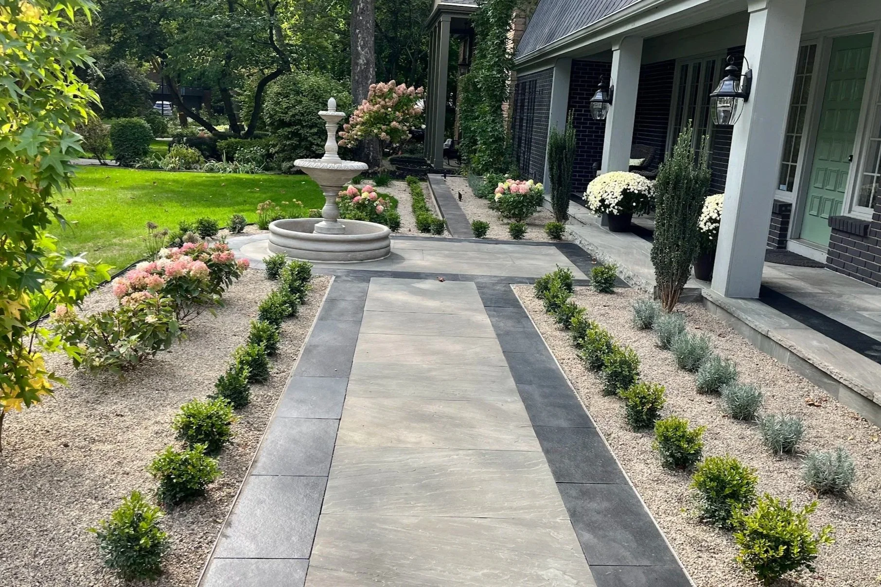 A landscaped front yard with a stone pathway leading to a fountain, surrounded by flower beds with pink and white flowers, and potted plants near the house entrance.