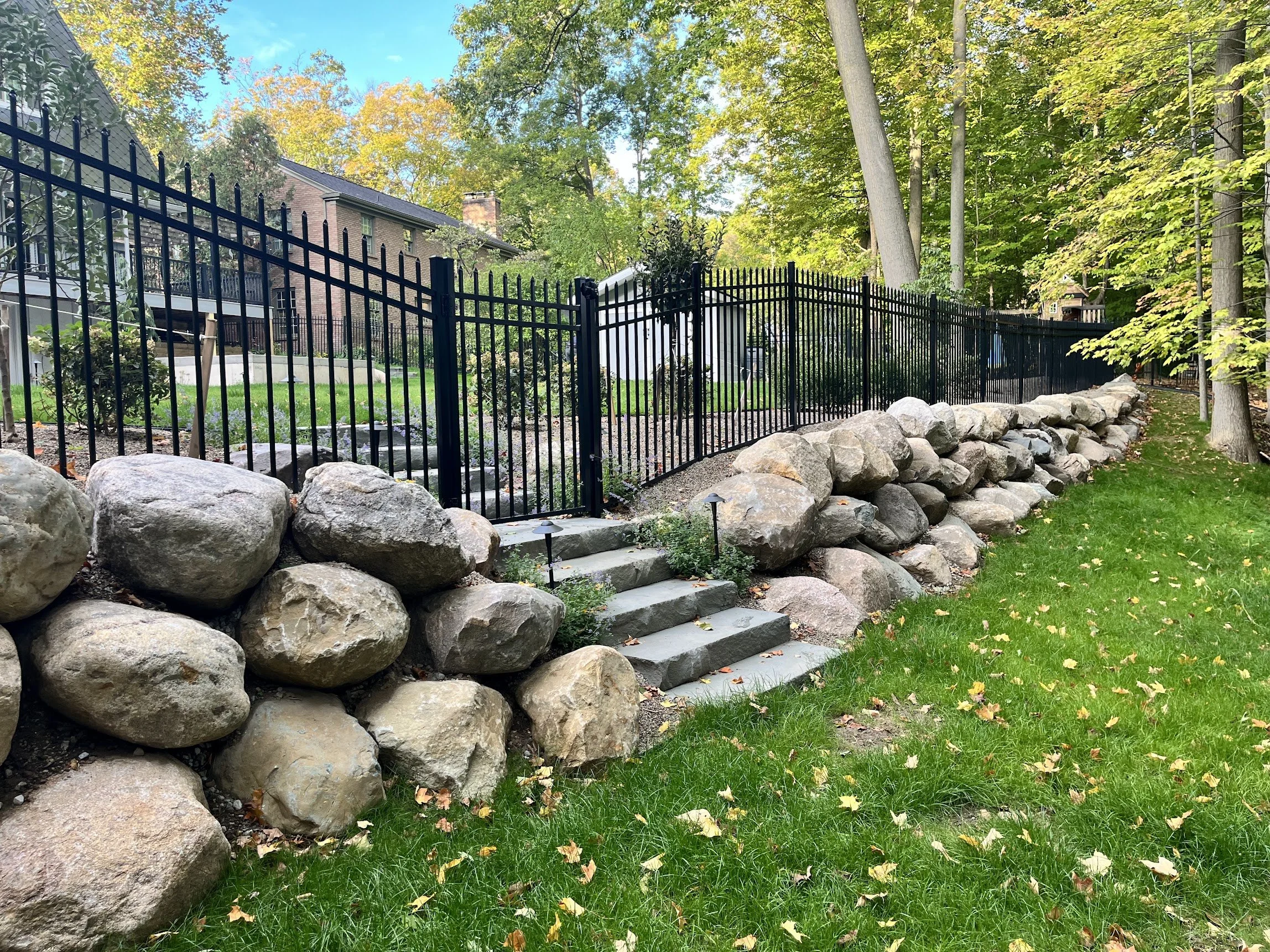 A landscaped backyard with a black metal fence, stone stairs leading up to the fence, large rocks lining the base of the stairs, and a grassy area with fallen leaves. There are trees and houses visible in the background.