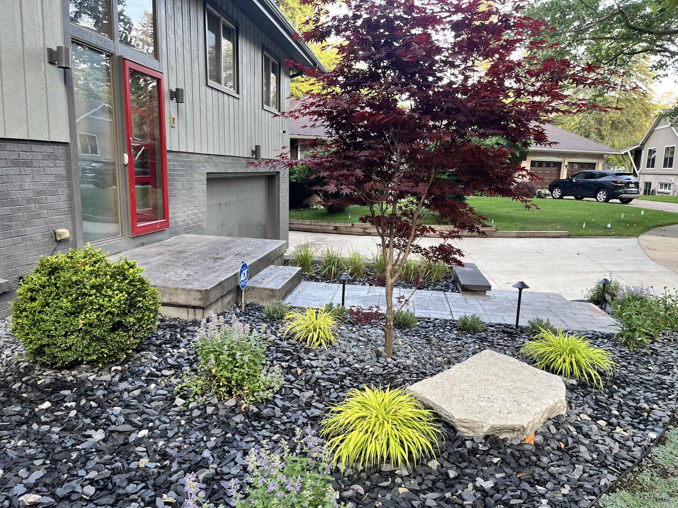 Front yard landscape with small red-leafed tree, various green plants, rocks, and a concrete porch.