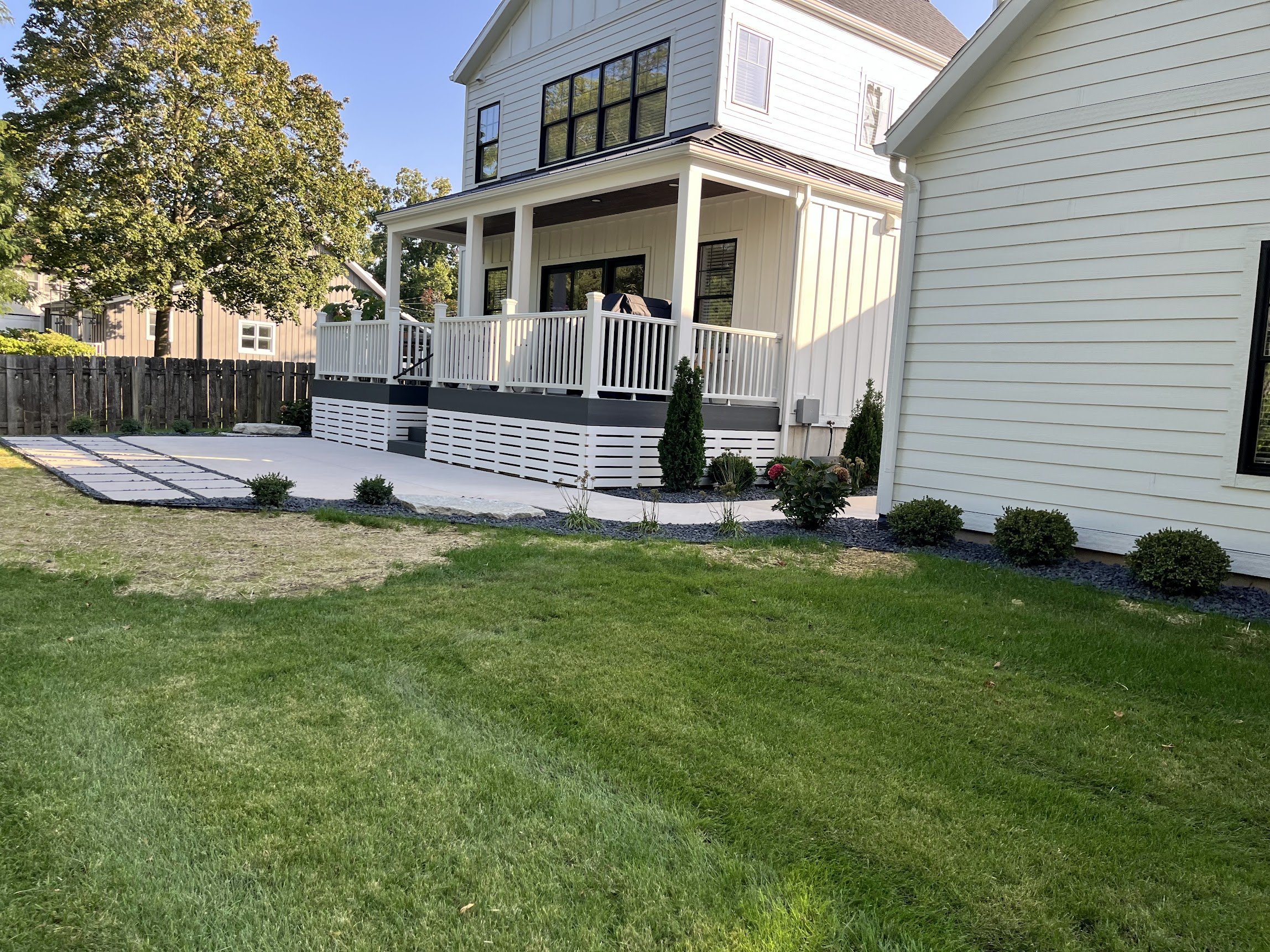 View of a backyard with a green lawn, landscaped flower beds, a wooden fence, and a white multi-story house with a porch and black window frames.
