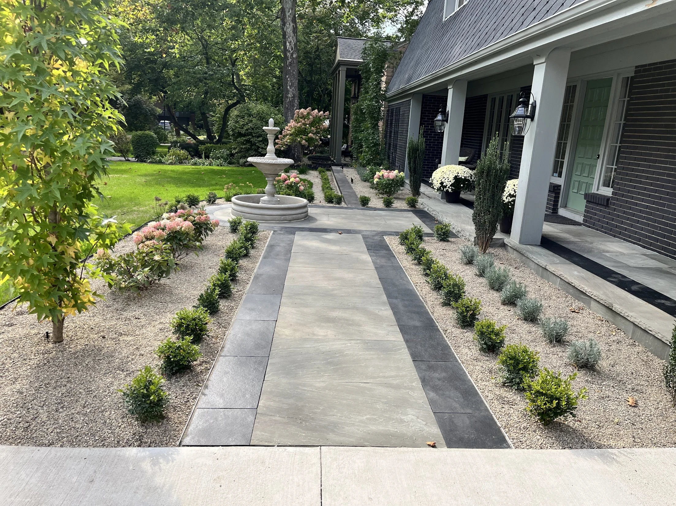 A well-maintained front yard with a paved walkway leading to a house, surrounded by flowering bushes, small plants, and a white decorative fountain, with trees and green grass in the background.