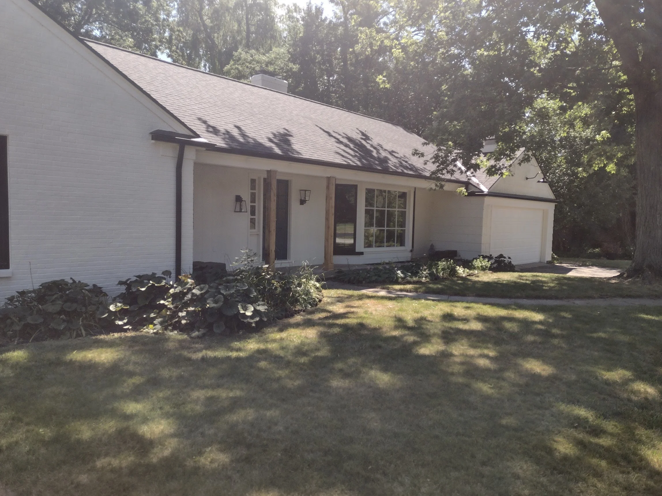 White house with a gray roof, large front window, front door, and attached garage, surrounded by greenery and trees, in sunlight.