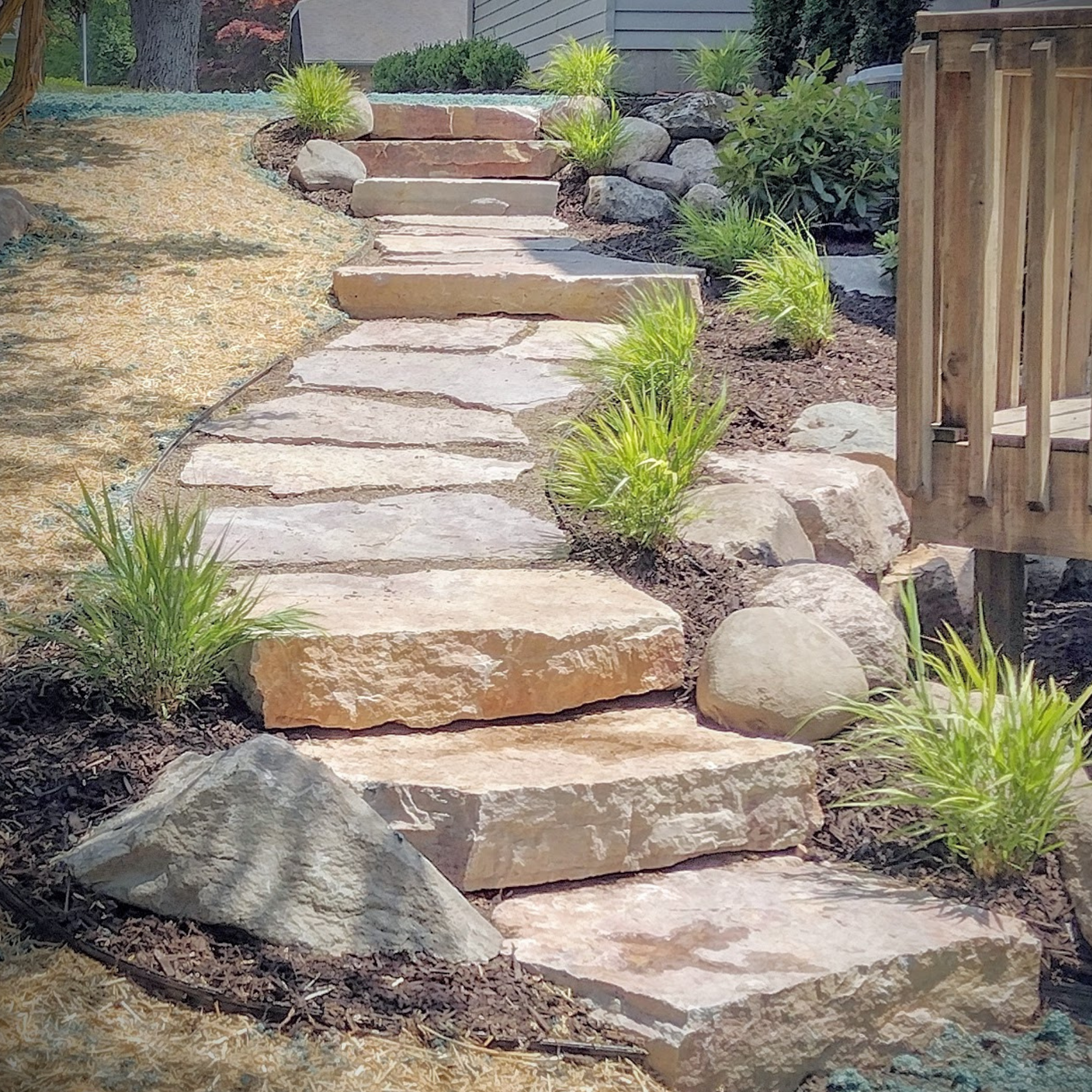 A stone pathway with large flat rocks and smaller round stones, bordered by green plants and shrubs, leading up a slight incline next to a wooden fence.