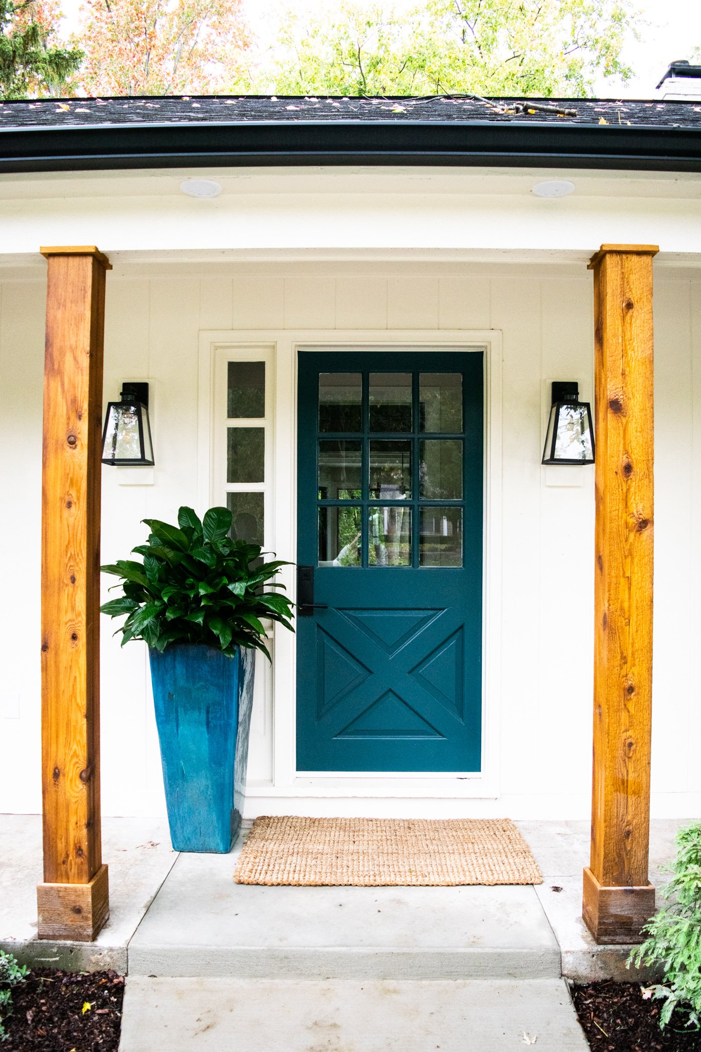 Front porch of a house with a blue door, wooden columns, two wall-mounted lantern lights, a potted plant, and a doormat.