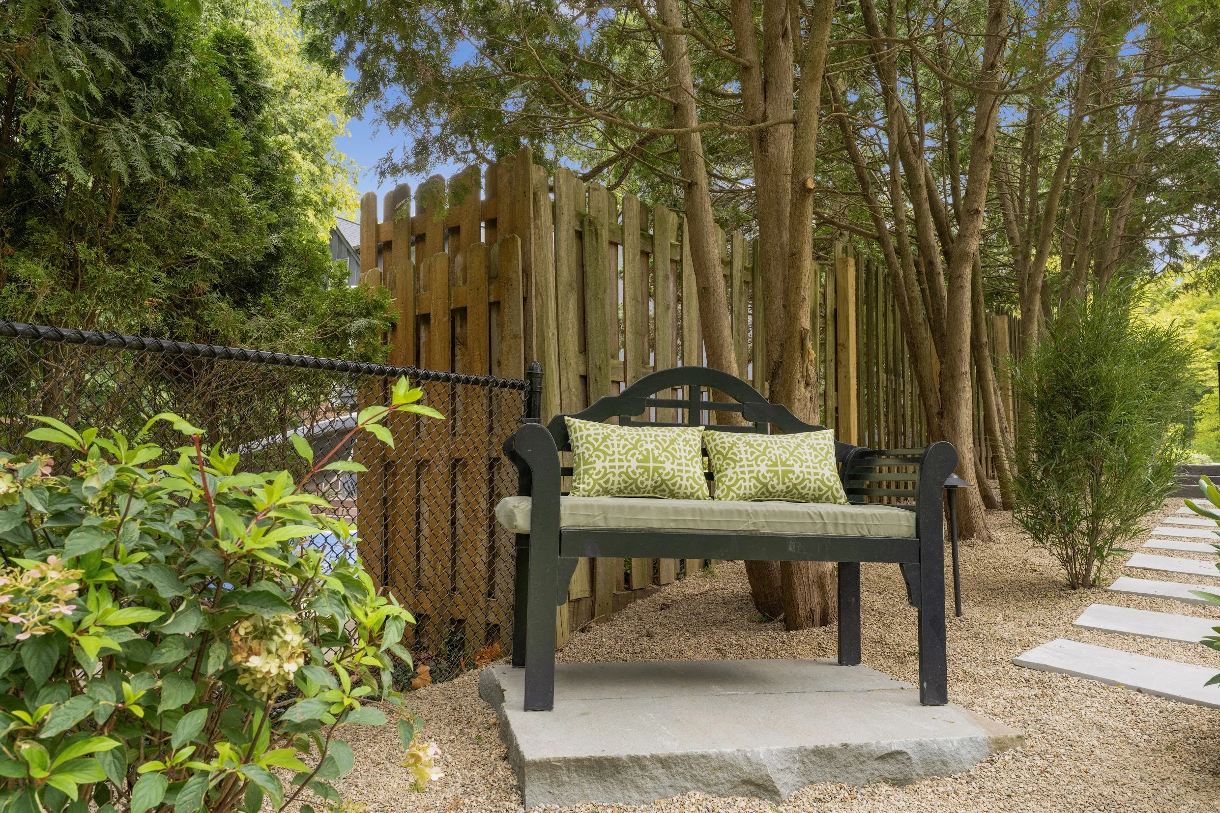 A black garden bench with two green patterned cushions sits on a concrete slab in a backyard, surrounded by trees, bushes, and a wooden fence.