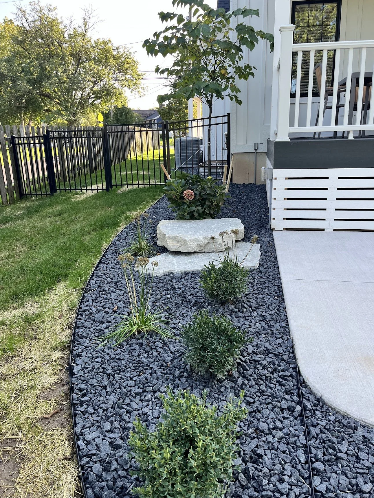Backyard garden with black gravel mulch, potted plants, and a small tree next to a house with a porch.