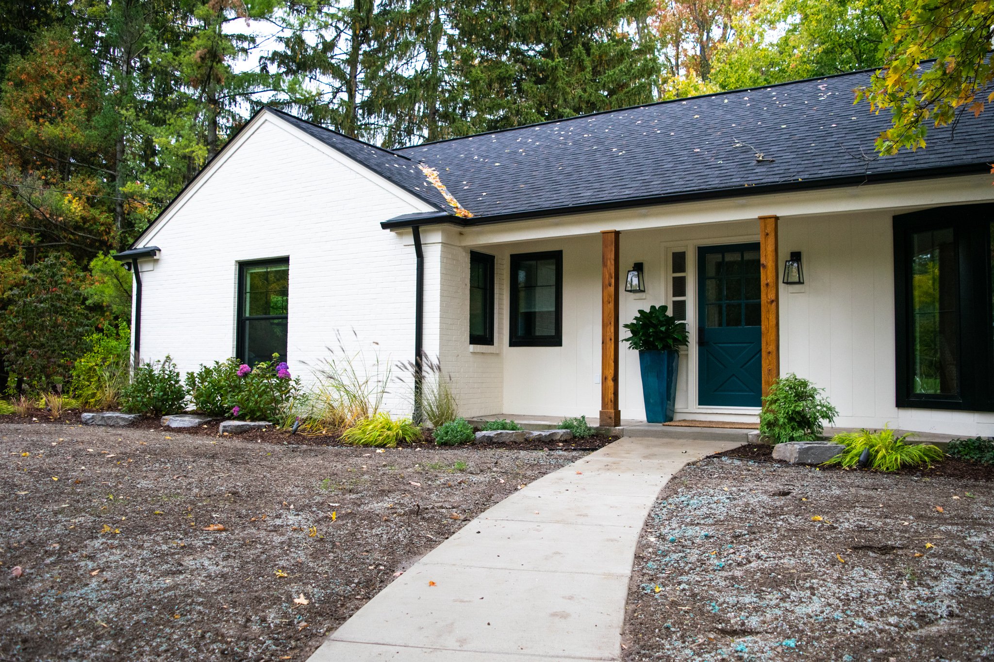 Front view of a modern house with white exterior walls, black framed windows, and a blue front door. The house has a black shingle roof and a small porch with wooden posts. There is a curved concrete walkway leading to the front door, surrounded by l